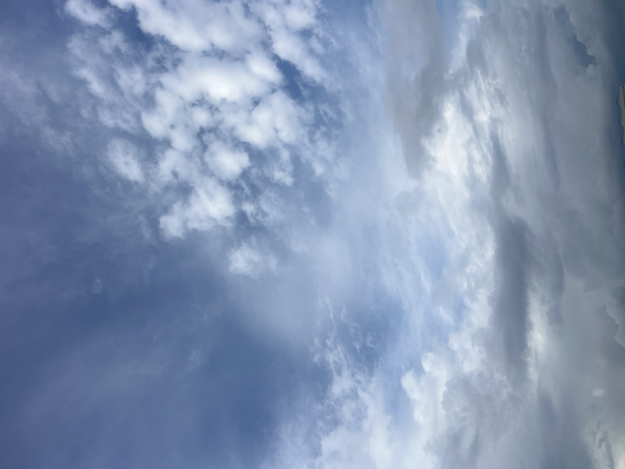 This media file depicts a sky filled with a mix of white and grey clouds, with patches of blue sky visible. The clouds are varied in texture and form, with some appearing wispy and scattered, while others are more dense and layered, suggesting a dynamic weather system. A sliver of a tall, modern building is visible on the far right edge of the frame, indicating an urban or semi-urban environment. The lighting suggests it is daytime, possibly late afternoon or early evening, given the presence of both bright and shadowed clouds. The overall scene is one of natural atmospheric beauty, with the weather appearing unsettled but not overtly stormy. There is no visible text in the image.