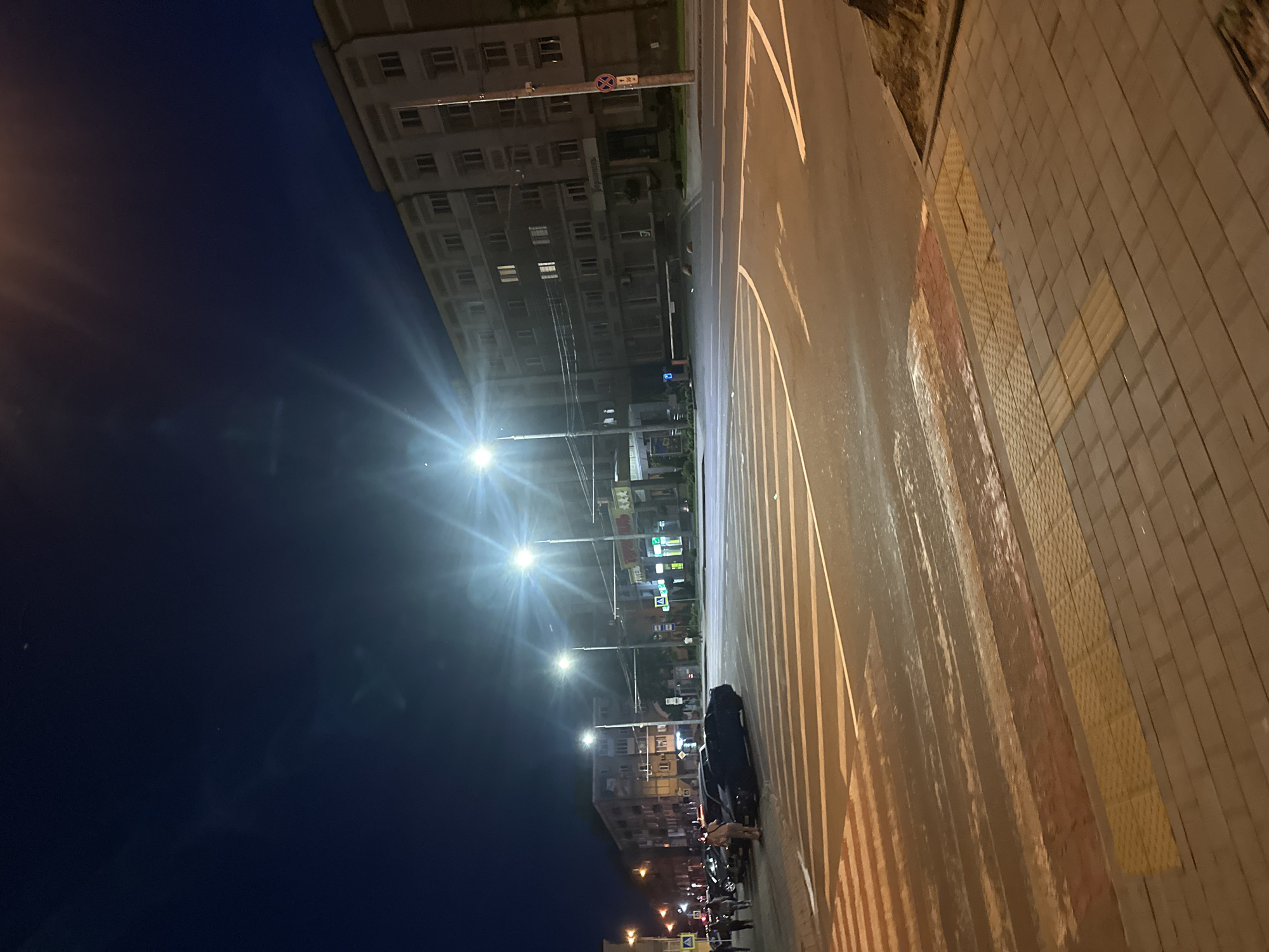 This image captures a nighttime urban street scene at a busy intersection in Ternopil, Ukraine. The specific location is identified as the intersection of Ruska Street and Hrushevskoho Street, facing generally west-northwest towards Ruska Street.
The scene is dominated by artificial lighting from numerous streetlights, casting bright light and prominent lens flares across the frame. The sky is a dark, deep blue, indicating either late evening or early morning.
In the foreground, a wide asphalt road with multiple lanes features a prominent pedestrian zebra crossing with white stripes, which appear somewhat reflective, suggesting a wet surface. Yellow tactile paving for the visually impaired is integrated into the sidewalk in the bottom right, which is constructed from rectangular and square paving stones.
On the right side of the image, a large, multi-story (approximately six floors visible) building with a light-colored facade (possibly beige or gray) stands prominently. This building features numerous uniformly spaced windows, several of which are illuminated, and some balconies. The ground floor clearly displays Cyrillic signage for "Укрпошта" (Ukrposhta - Ukrainian Post Office), and a red circular "no stopping" or "no parking" sign is affixed to a pole in front of it. A white barrier gate is visible across a paved entrance to the building's side or rear. Further up on the same building, another sign displaying "ГОТЕЛЬ" (HOTEL) is visible, along with another commercial sign.
On the left side of the street, further in the background, several other multi-story residential or commercial buildings are visible, also illuminated. A group of several pedestrians is observed on the sidewalk on the left, near a row of parked cars. At least one dark-colored sedan is clearly visible parked next to the sidewalk. Another individual is seen walking on the sidewalk near the main building on the right.
Street furniture includes multiple tall lamp posts equipped with bright white light sources. Blue rectangular road signs with white arrows, indicating traffic flow or turns, are also present. Sparse vegetation, possibly trees or bushes, lines parts of the sidewalks. The overall environment suggests a well-lit, active urban area at night.