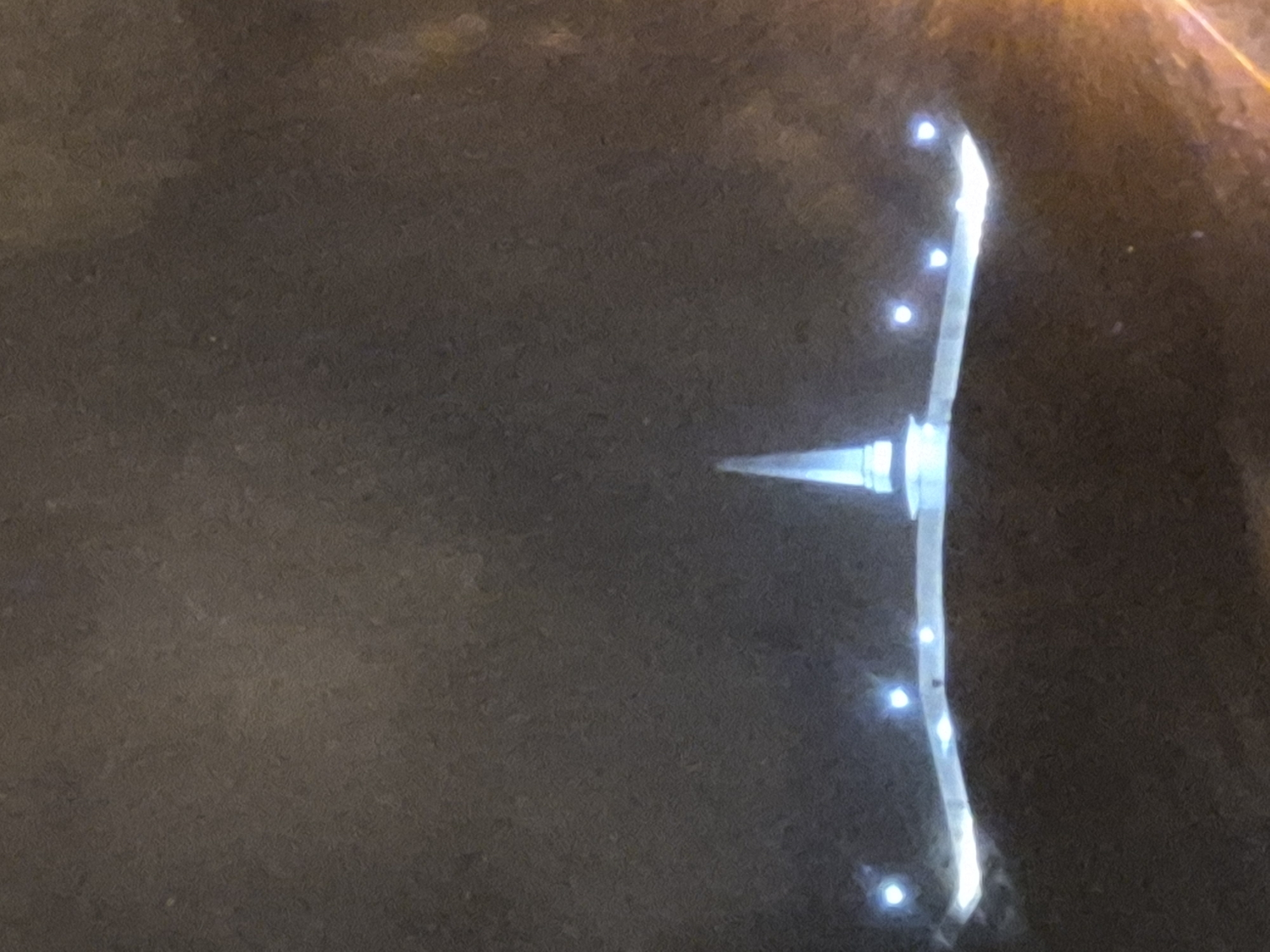 The image displays an abstract, illuminated structure against a dark, textured background at night in Tobolsk, Russia. The primary subject is a modern-looking installation featuring a central, sharp, conical element emitting a bright white-blue light. This central piece is integrated into a longer, curved, and highly reflective or light-emitting arm. Along this curved arm, several small, distinct blue-white point lights are regularly spaced, resembling embedded LED lights. The surrounding environment is dark, suggesting nighttime, with a somewhat granular or uneven surface that could be ground or paving. No people or specific activities are visible, and there is no discernible text. The overall impression is one of modern artificial lighting in a dark urban or outdoor setting.