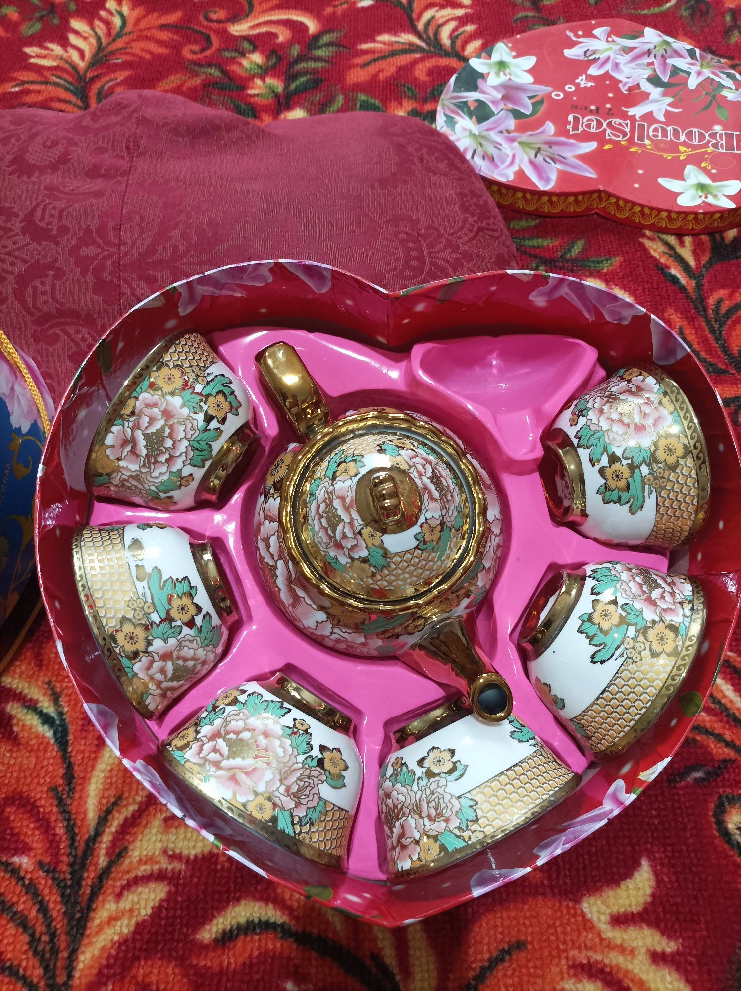 In Seýdi, Turkmenistan, a heart-shaped red box contains a porcelain tea set with floral and gold accents. The set includes a teapot and six small cups, all intricately decorated with pink and white peonies and gold geometric patterns. The box is placed on a red fabric with a vibrant, abstract floral design. A lid for the box, also red and decorated with lilies, is visible to the right of the main image. The scene appears to be indoors, likely in a home setting, given the fabric background. There are no people or actions depicted in the image.