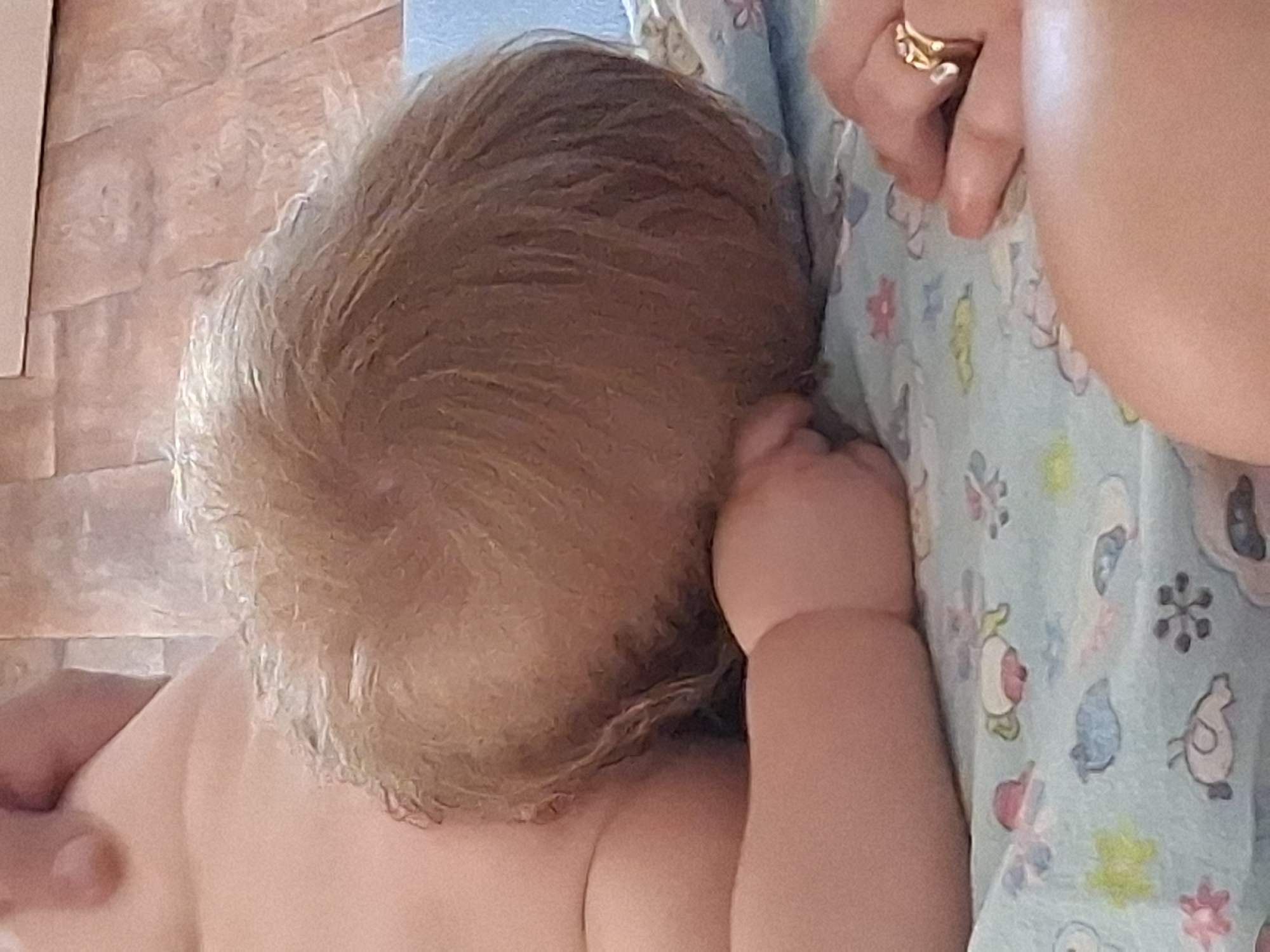 This media file features a close-up, overhead view of a baby's head, with fair hair styled in a whorl. The baby is nestled against a person whose arm and shoulder are visible on the right side of the frame, and whose hand, adorned with a gold ring, is gently placed on the baby's head. The baby is lying on a light blue blanket patterned with cartoon animals and flowers, including what appear to be birds, bunnies, and other whimsical figures. To the left of the baby's head, a portion of a tiled wall or surface is visible, rendered in warm, earthy tones. The lighting suggests an indoor setting, possibly during the daytime. The overall mood conveyed is one of tenderness and care, focusing on the intimate connection between the baby and the caregiver. No text is visible in the image. The provided location context is Semey, Kazakhstan, but this information cannot be directly inferred from the visual content of the image itself.