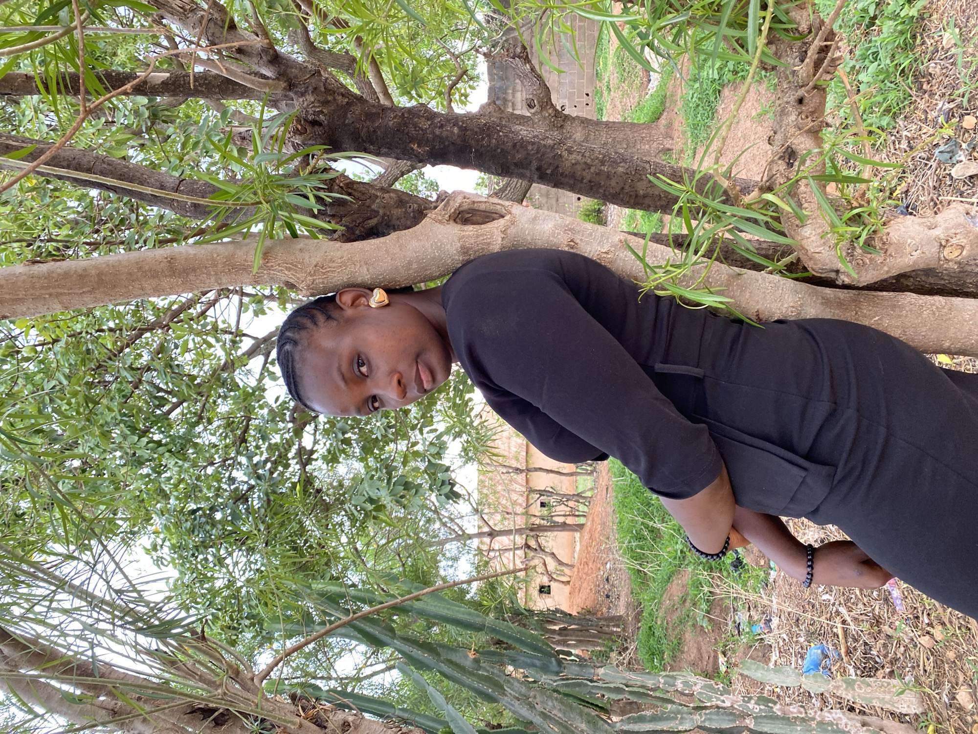 A woman leans against a tree in Kuru Dungus, Nigeria. She wears a black outfit and a gold earring. Her hair is styled in cornrows. She is positioned to the left of the frame with her head close to the tree trunk. The background shows greenery, a building, and other trees, suggesting a natural environment near the edge of the city.