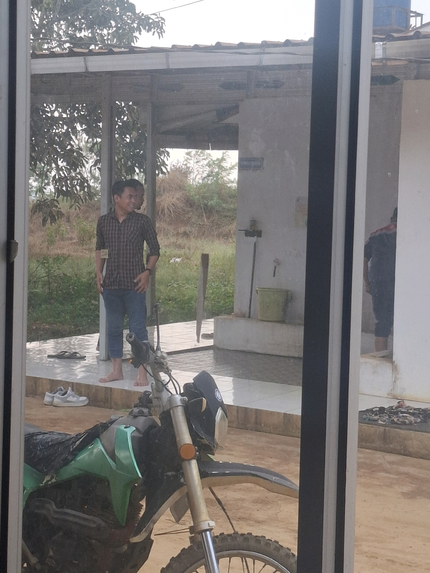 A man wearing a plaid shirt and blue pants stands on a tiled porch. He is barefoot. To his left, on the porch, are a pair of dark sandals. Further to the left, on the ground, are a pair of white sneakers. A green motorcycle is parked in the foreground. The man is smiling and looking to his right, with another person's silhouette visible behind him. In the background, there is a building with a corrugated metal roof, trees, and dry vegetation. A green bucket and a faucet are mounted on a wall to the right of the man. The scene appears to be outdoors in a rural or semi-rural setting.