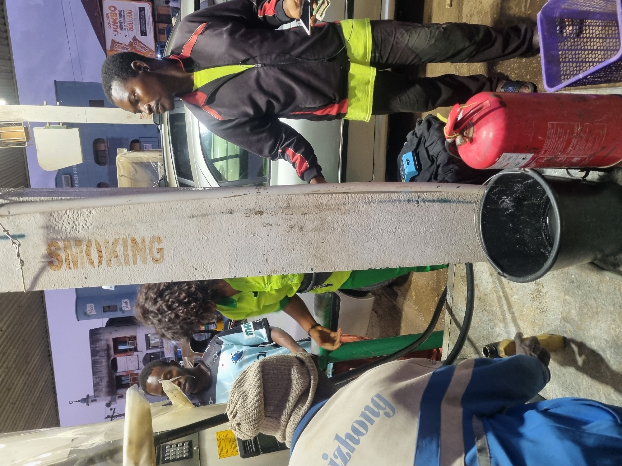 Here's a detailed description of the image:
**Setting:** The image appears to be taken at a petrol station in Jos, Nigeria. Evidence for this includes:
* The presence of petrol pumps and fuel dispensing equipment.
* Signage bearing the name "Golden Jumbo" on a building behind the station, which may be the name of a product sold here.
**People:** The image features three individuals.
* One is standing. This person is wearing a dark jacket with neon yellow panels and red accents. They are holding a phone and cash, potentially making a payment.
* A second individual is assisting with fueling. This person is wearing a light blue and white shirt with the word "MAL" or "MAT" visible on the chest. They also wear a safety vest.
* A third individual is also present wearing a blue and white jacket with the letters 'zhong' on it and a light gray knit cap.
**Objects:** Key objects in the scene include:
* A petrol pump with a fuel nozzle in use.
* A red fire extinguisher sitting on top of a small black travel bag.
* A black bucket.
* A purple plastic basket
**Actions:**
* A gas station attendant fills up a vehicle.
* Another worker is handling payment.
**Environment:** The scene appears to be in an urban area with buildings visible in the background. The lighting suggests that it may be evening or night. A sign warning of "Smoking" is prominently displayed near the pumps.