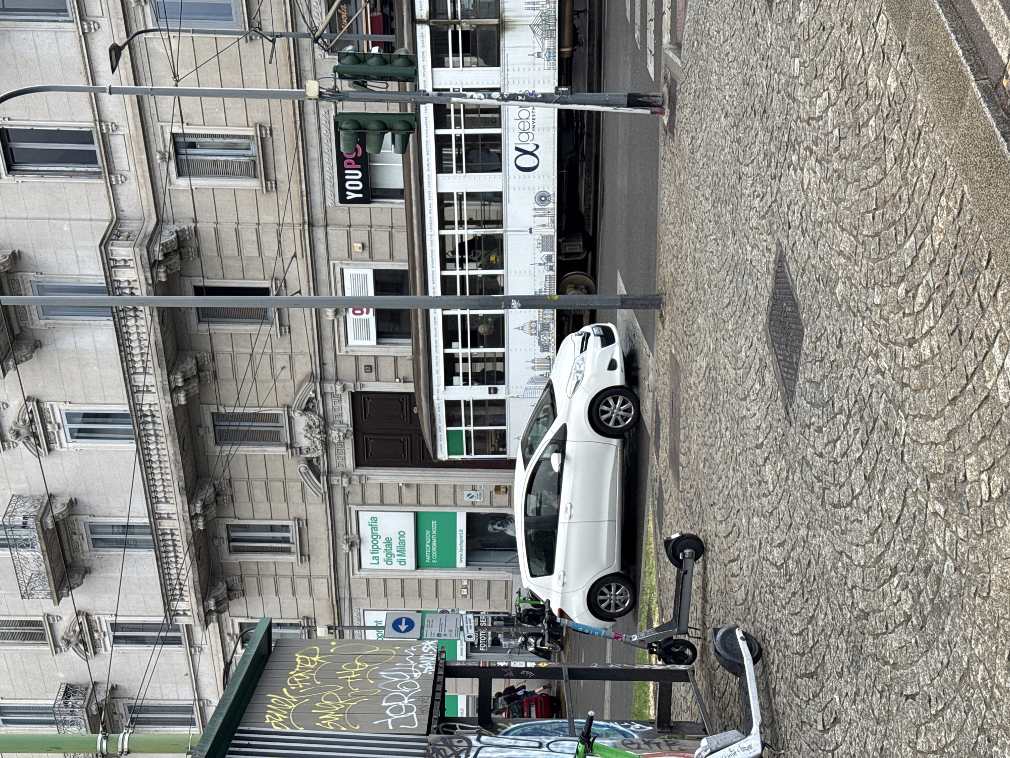 In Milan, Italy, a street scene unfolds with a white hatchback car and a tram as the primary subjects. The car, a Toyota Yaris, is parked on the left side of the street, facing right. The tram, adorned with drawings of landmarks from various global cities, is on the right, traveling left. A person is visible inside the car, looking out the window. The street is paved with cobblestones, and a light pole with traffic signals stands in the foreground. A street art-covered structure is on the far left, with two electric scooters parked in front of it. In the background, a multi-story building with balconies and windows stands tall. The sky is overcast.
