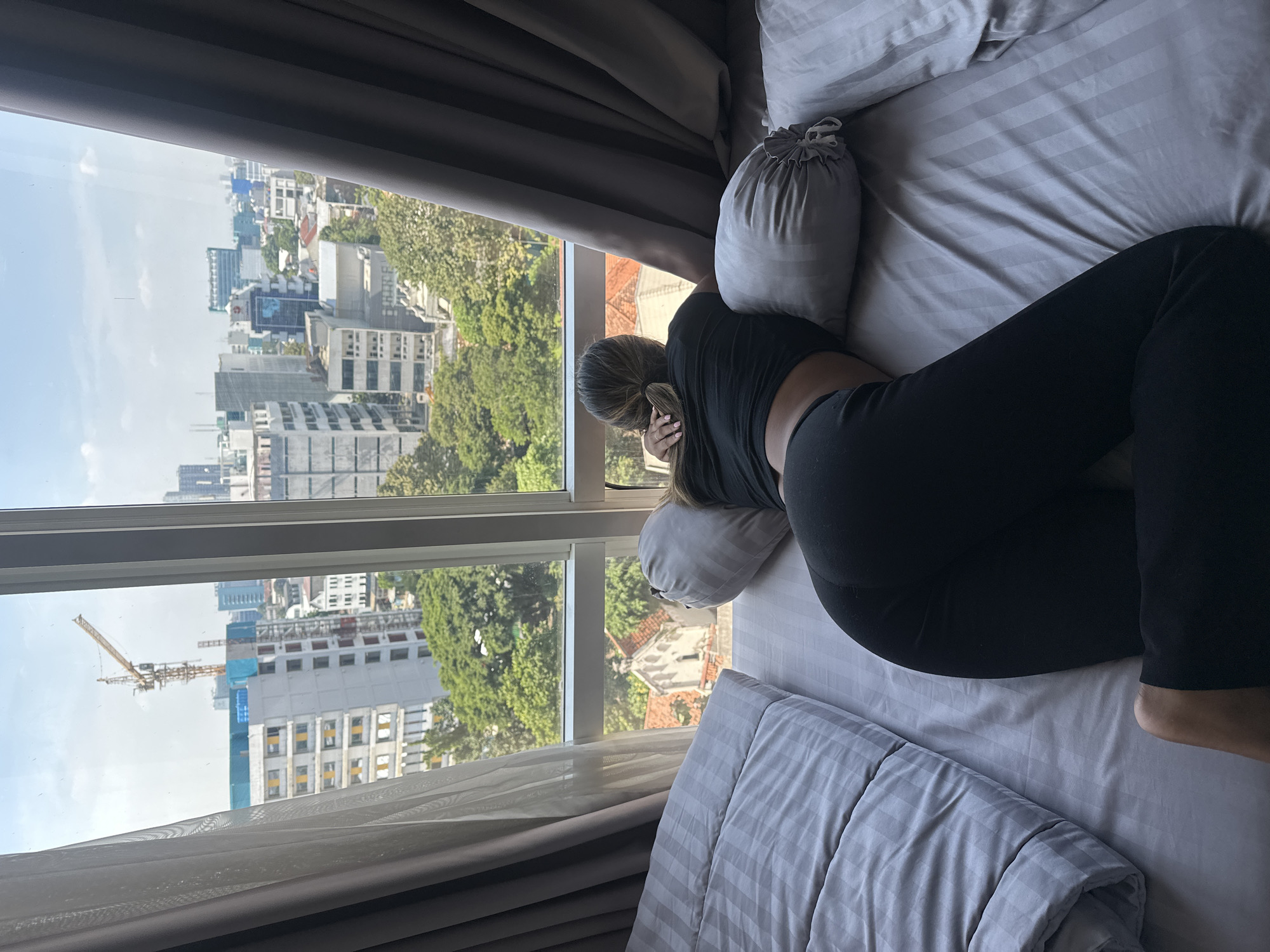 A woman lies prone on a bed facing a large window in what appears to be a hotel room in Central Jakarta, Indonesia. The city skyline, with trees interspersed among the buildings, is visible through the window. A construction crane is visible in the lower left. The woman is wearing dark clothing and is resting her head on a pillow. Gray and white striped bedding is visible. Curtains frame the window.