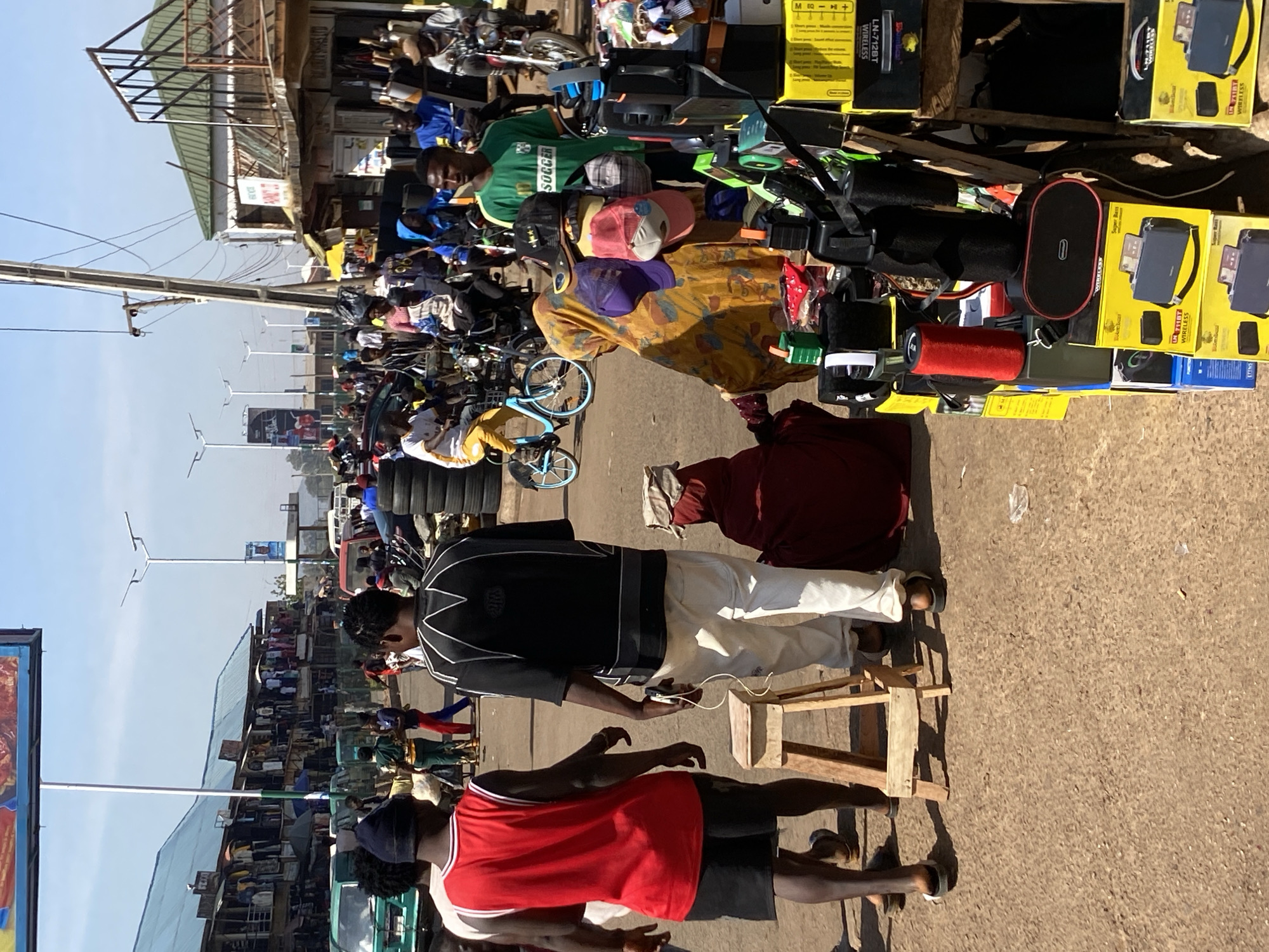 A ground-level, daytime shot depicts a bustling street scene in Bukuru, Nigeria. The foreground shows two individuals, one in a red basketball jersey and the other in a black and white shirt, standing near a small wooden stool. The latter appears to be using a mobile phone. Behind them, a vibrant marketplace is teeming with people and diverse goods. The array of items includes speakers, hats, and other merchandise. The street is lined with shops and buildings, some with corrugated metal roofs. In the distance, utility poles and streetlights punctuate the urban landscape. The sky is clear.