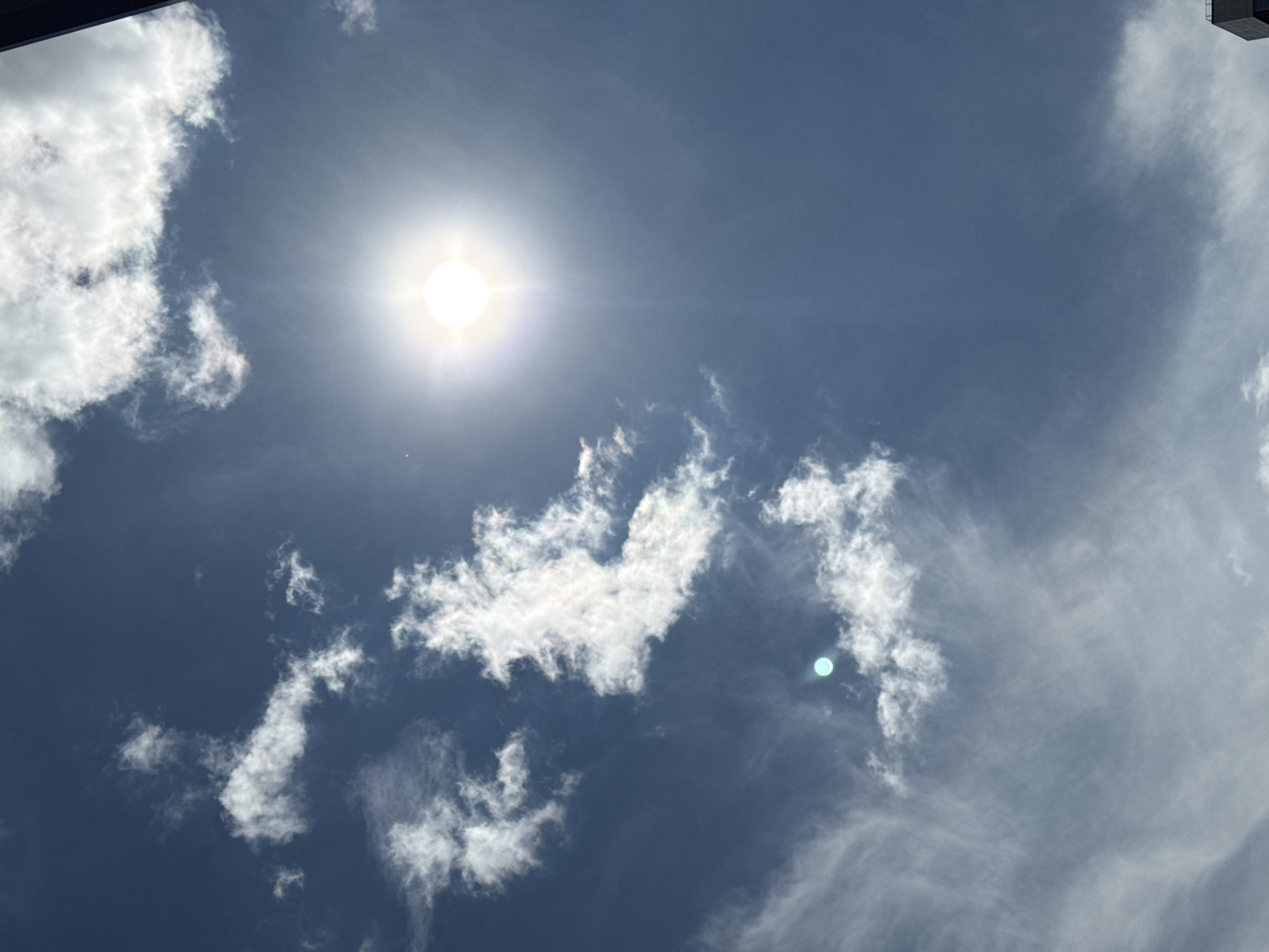 A bright, sunny sky with scattered clouds is visible. The sun is prominently featured in the upper right quadrant, emitting a strong glare. Several white, fluffy clouds are dispersed across the blue sky, some appearing wispy and thin, while others are more substantial. In the lower right corner, the edge of a dark, rectangular building is visible, suggesting an urban or semi-urban setting. The scene is devoid of any people or discernible actions. This image is a view of the sky from Pargolovo, Russia, on a clear day.