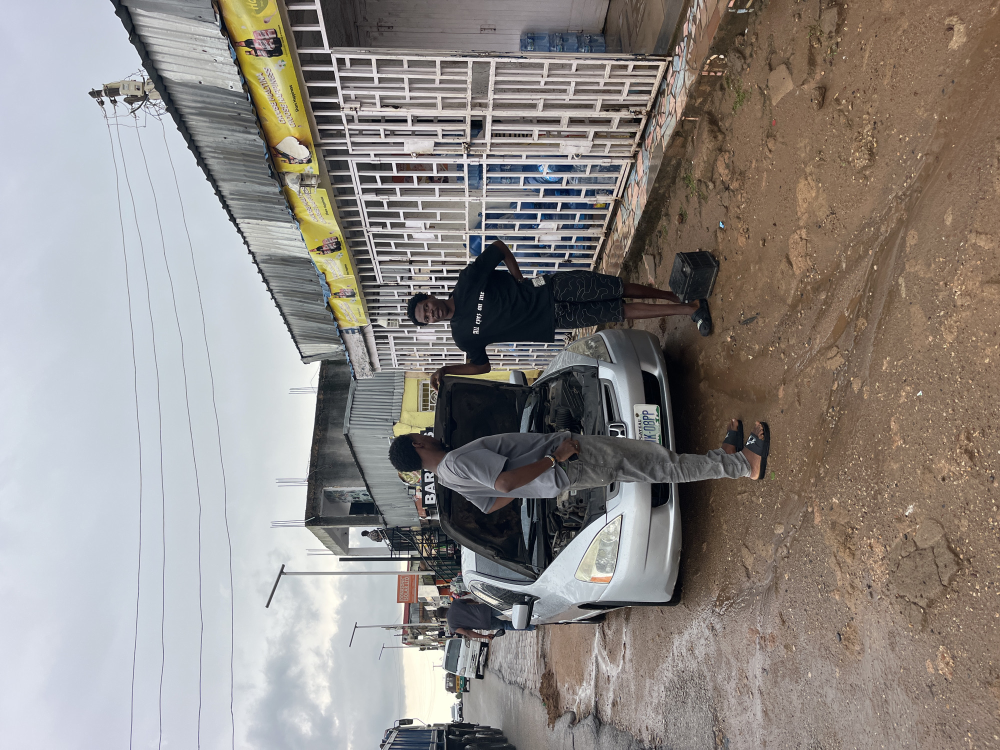 This image shows two young men working on a silver car with its hood open. One man, wearing a black t-shirt and patterned shorts, stands by the driver's side door, leaning against the car. The other man, in a grey t-shirt and grey pants, is looking into the engine bay. A black toolbox sits on the muddy ground to the right of the car. In the background, there's a building with a white metal grate in front of it, and a corrugated metal roof overhang with yellow banners. One banner displays the text "GHOUSEMALTINA CHOOSE HAPPINESS Share Happiness". Another sign on the building reads "DONASO SOLAR LTD BAR". The ground is wet and muddy, suggesting recent rain or a damp environment. The sky appears overcast. The overall scene appears to be an outdoor repair shop or a roadside service area in an urban or semi-urban setting. The license plate of the car is visible and reads "PLATEAU K-08PP". The man in the black t-shirt has text on his shirt that reads "All eyes on me".