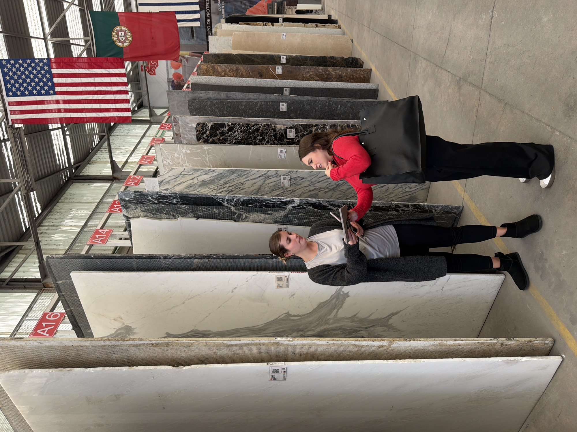 Two women are in a warehouse-like space examining large slabs of marble or granite, presumably for countertops or similar purposes. One woman wears a red long-sleeved shirt and black pants, carrying a large black tote bag. She is leaning forward, appearing to engage in conversation with the other woman. The second woman is wearing a white shirt under a dark gray cardigan sweater, with black leggings and shoes. She is standing on a slab of marble and holding a notebook.
The marble slabs are displayed in vertical racks, with labels. The background shows various types of slabs, some with dark veining and others with lighter colors. American and Portuguese flags are seen hanging from the ceiling in the background, suggesting international sourcing of the materials.
The location is implied by an image search to be somewhere in or around Puebla, Mexico, with the presence of both American and Portuguese flags alongside Spanish-language material suggesting the location.