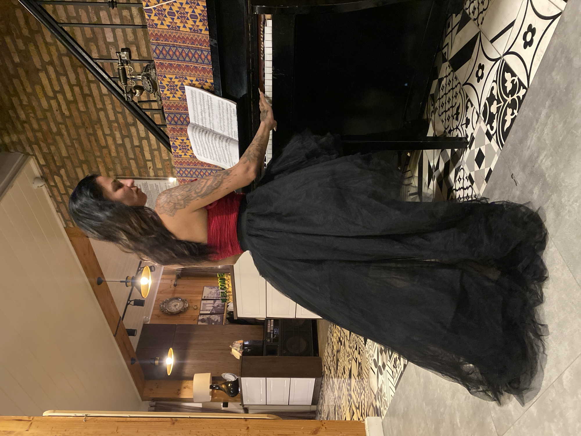 A person is playing a piano in a room with wooden floors and brick walls. The person has long, dark hair and is wearing a red top and a long black skirt. The piano is open and sheet music is on the stand. The room is decorated with a colorful rug, a vintage telephone on the wall, and several framed pictures. There are also two lamps with yellow light illuminating the space. The floor has a black and white geometric tile pattern. The overall mood is serene and focused.