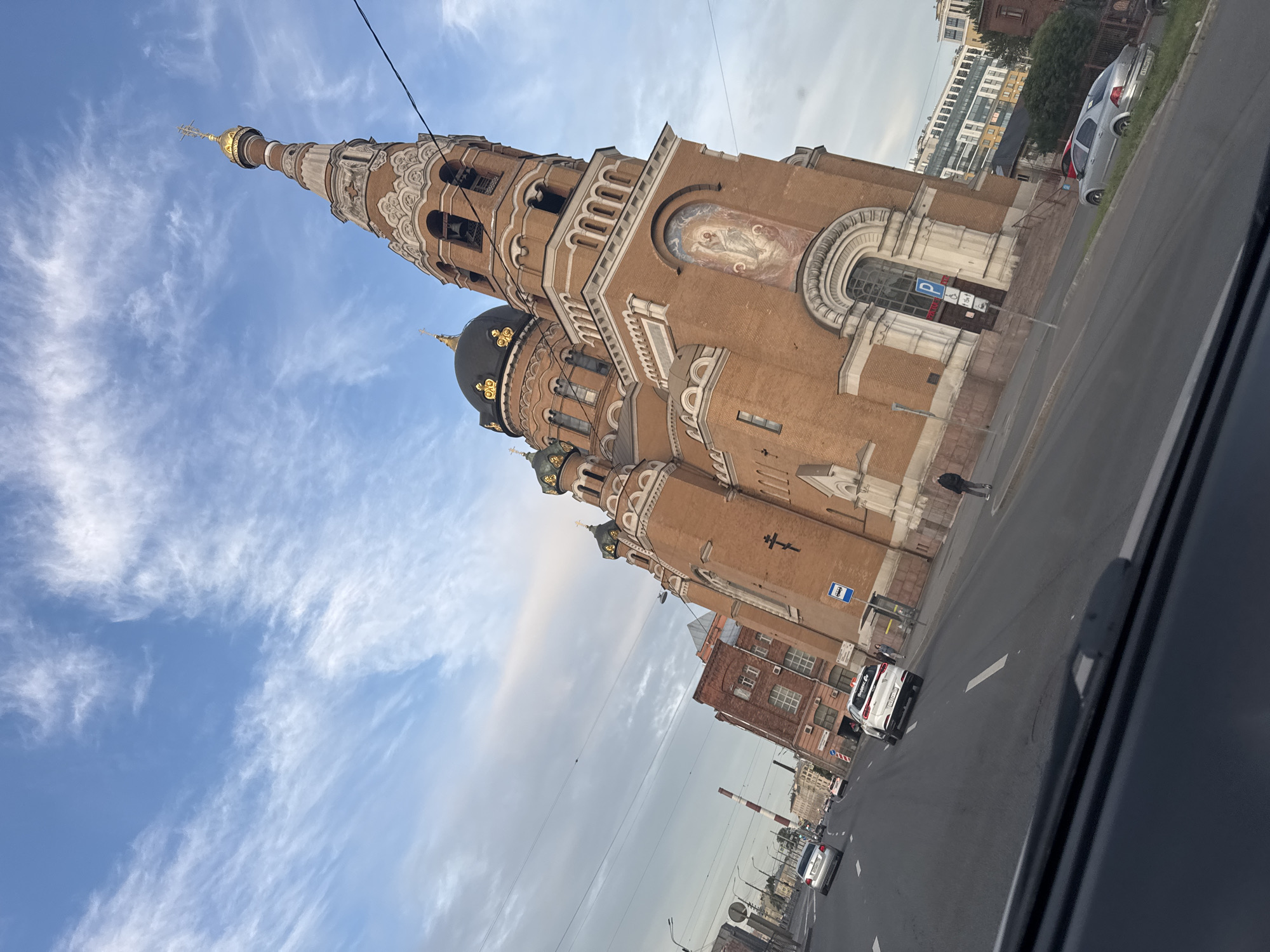 The image depicts a view from inside a vehicle, looking out at a large, brick Orthodox church. The church has multiple domes, including a prominent black dome with gold accents and a tall, slender spire topped with a golden cross. The architecture features intricate detailing, such as arched windows and decorative friezes. A religious mural is visible on one of the church's walls.
The scene is set on a street next to the church, with a road and several cars visible. One car, a white vehicle, is parked on the side of the road. Another white car, possibly a taxi, is driving away from the camera. In the distance, other buildings and a railway crossing signal can be seen. The sky is blue with wispy white clouds, suggesting it is daytime. The overall mood of the image is calm and observational.
There is some visible text in the image. On the white car driving away, there is text that appears to read "Yandex Go". A blue sign with a white "P" indicating parking is also visible on the side of the street. Another blue sign with white text and symbols, possibly related to public transport or a bus stop, is also present. The photo was taken in Saint Petersburg, Russia.