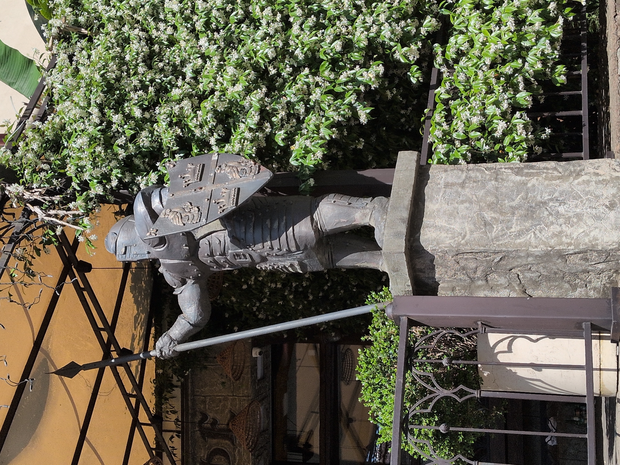 The image shows a life-sized statue of a knight in full armor holding a spear and a shield. The statue is positioned outdoors, in front of a building with a textured facade and a large window. It is surrounded by lush greenery, including a dense wall of flowering plants and a potted plant behind a decorative metal fence. A yellow awning is visible overhead. The scene appears to be a sunny day. The specific location in the city of Sochi, Russia, is not discernible from the image itself.