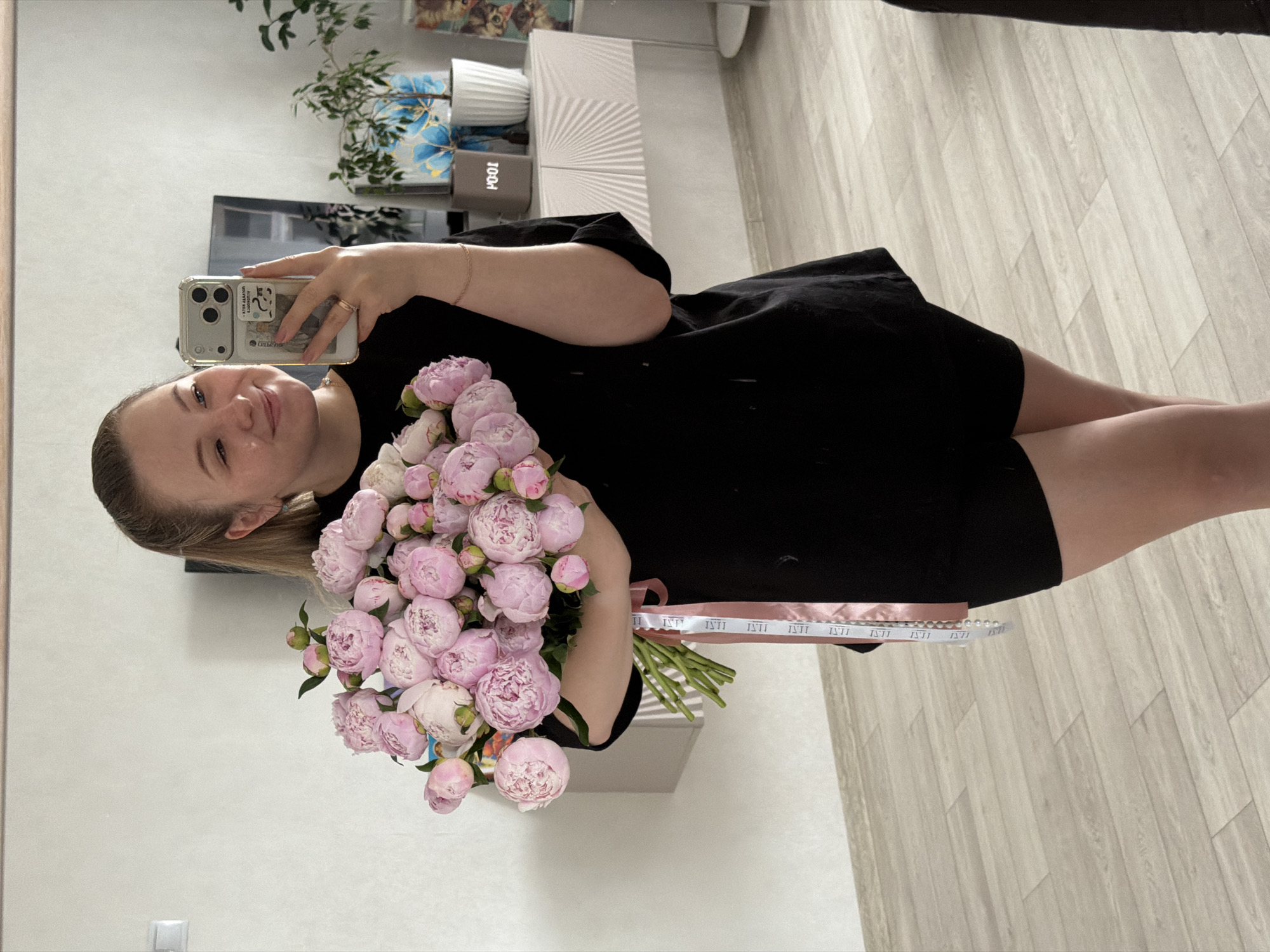 This image captures a full-body mirror selfie of a young adult female indoors, located in what is assumed to be the city of Chaikovsky, Russia. The female, with light-colored hair pulled back, stands facing the mirror with a slight smile. She is wearing an oversized black t-shirt and black shorts. On her right hand, she wears a ring, and a thin, light-colored bracelet is visible on her left wrist.
She holds a large bouquet of light pink peonies in her left arm, with the green stems visible. The stems are secured by two ribbons: one solid light pink and another white ribbon patterned with repeating "ISTI" text in black. A string of white beads or pearls also hangs from the ribbons. In her right hand, she holds a light-colored smartphone, enclosed in a clear protective case with a small dark sticker or charm on the back. The phone screen reflects a reversed image of the subject and the room.
The background, seen through the mirror reflection, features light-colored walls and a light-toned wooden or laminate floor laid in planks. To the viewer's left, a white light switch is mounted on the wall. To the viewer's right, a dark television screen is visible above a white console table or cabinet. This console table exhibits a textured, radial design on its front surfaces. Items on the console table include two potted plants, a decorative item with a blue and gold floral pattern, a digital clock or device displaying "10:01" (possibly reversed "PO:01"), and a partially visible framed image of cats. The room is illuminated by ambient light.