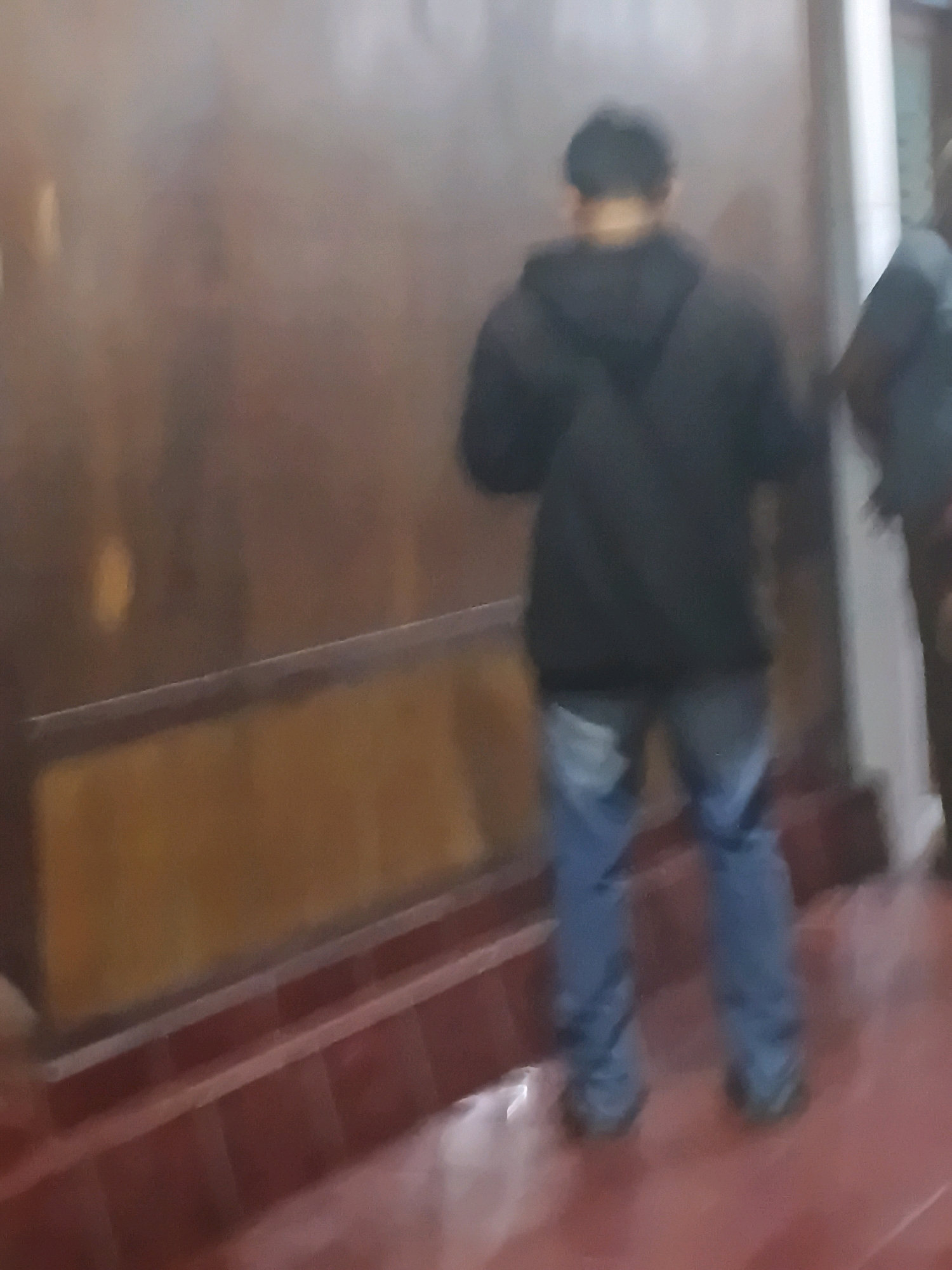 In the city of Purwodadi, Indonesia, a person stands with their back to the camera, facing a large, reflective surface that appears to be a wall or partition. The individual is wearing a black hooded jacket and blue jeans. The reflective surface has a reddish-brown hue and a horizontal band of gold-colored material is visible on its lower half. The floor in front of the person is a deep red, with lighter red lines suggesting a tiled pattern. To the right of the main subject, another person, partially visible and out of focus, appears to be standing. The overall lighting suggests an indoor environment.