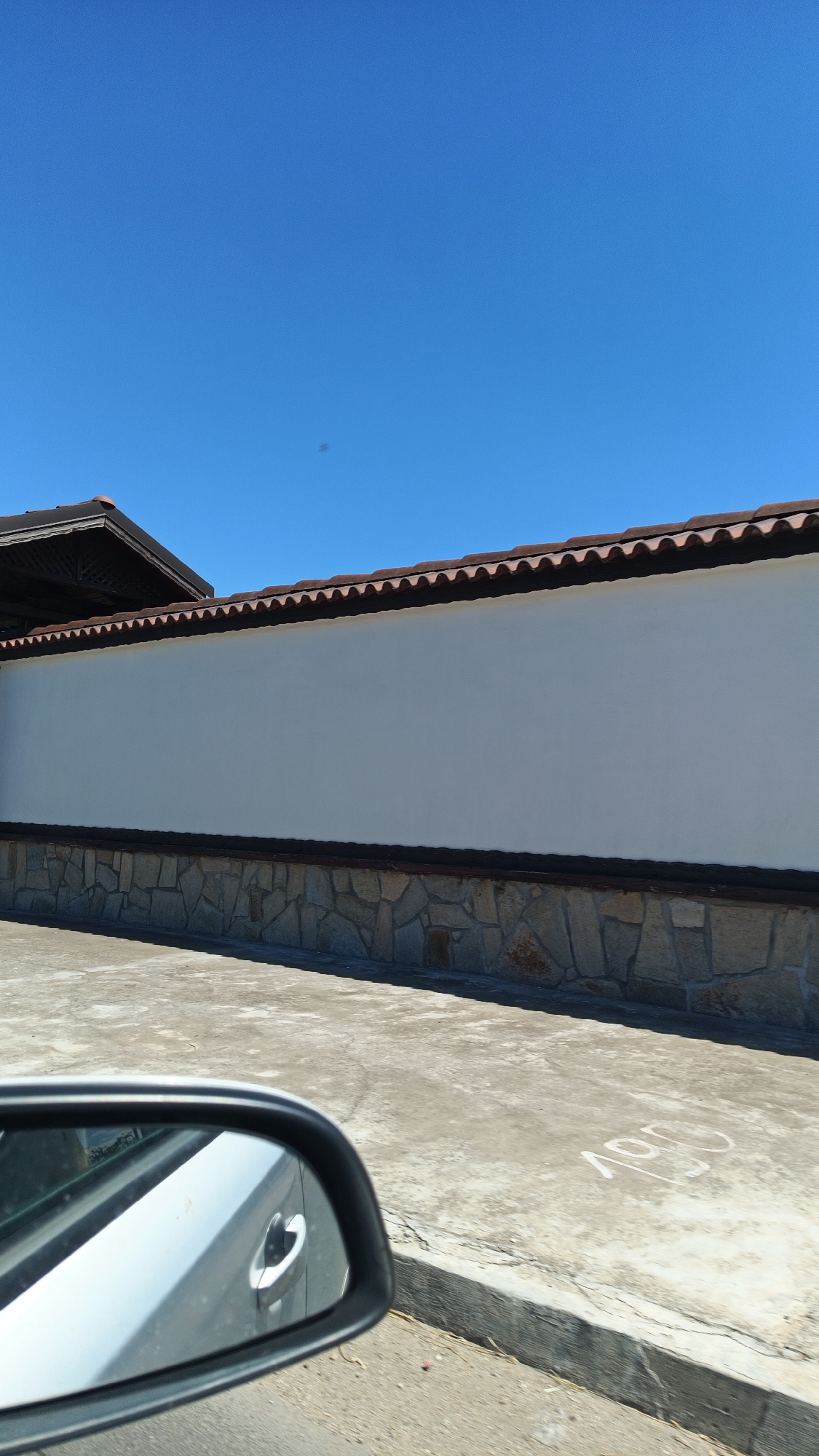 The image shows a building facade with a white wall, a stone base, and a tiled roofline under a clear blue sky. The facade is situated along a concrete area with markings, potentially a parking lot. A car's side mirror is visible in the foreground. The location is assumed to be Milevo, Bulgaria.
