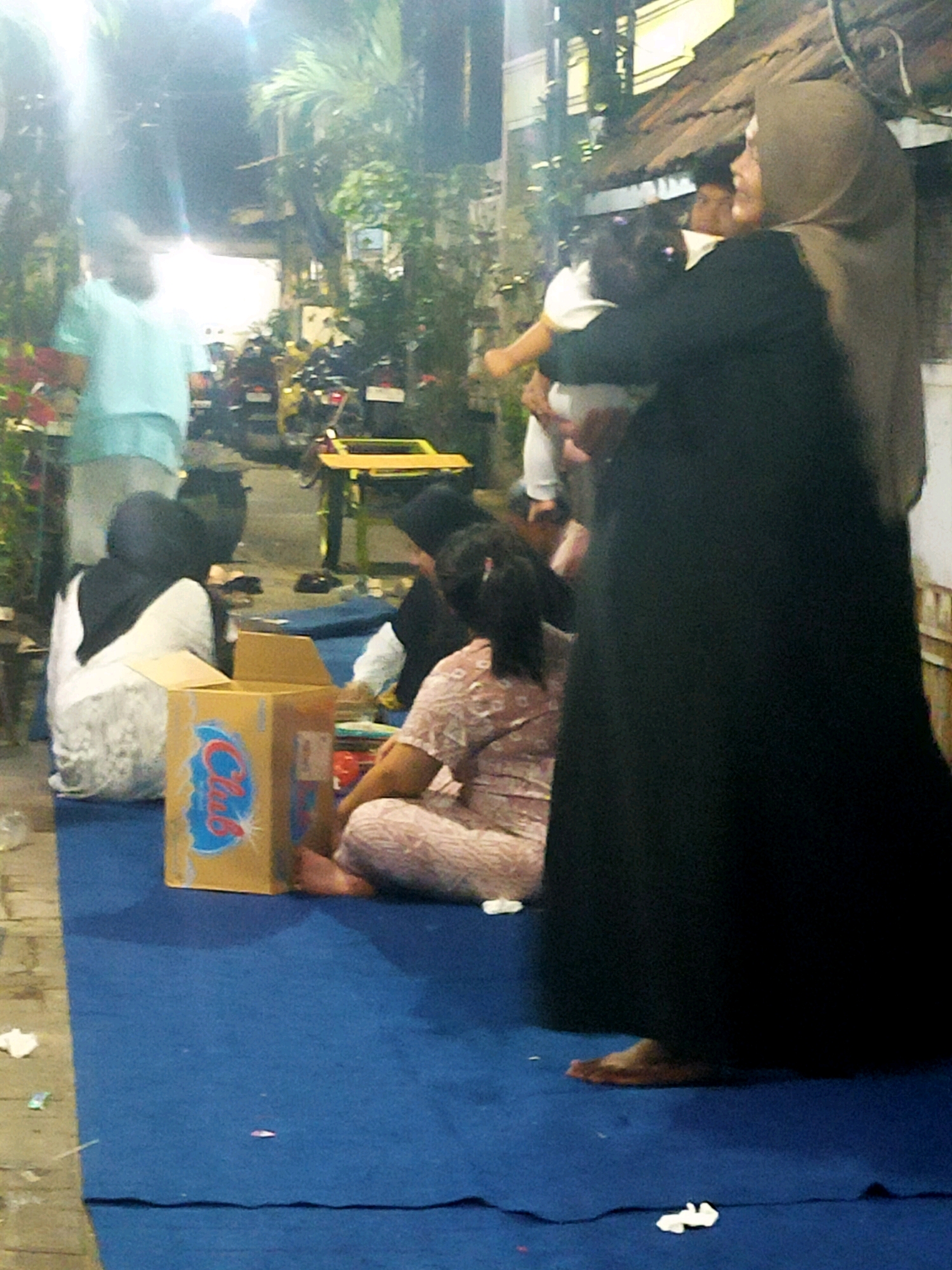 The image depicts a nighttime street scene in Surabaya, Indonesia. Several people, including women and children, are gathered on a blue mat placed on a paved surface. One woman in the foreground wears a black abaya and a light brown hijab, holding a child. To her left, another woman in a white hijab and black attire is seated. Further in the background, a man in a light blue shirt and a child in white are visible. A yellow table with a green base is set up behind the seated individuals, and several motorcycles are parked in the background. A cardboard box with a "Club" logo is placed on the blue mat. The environment appears to be a street or alleyway with overhead lighting casting shadows and creating a somewhat blurry atmosphere.