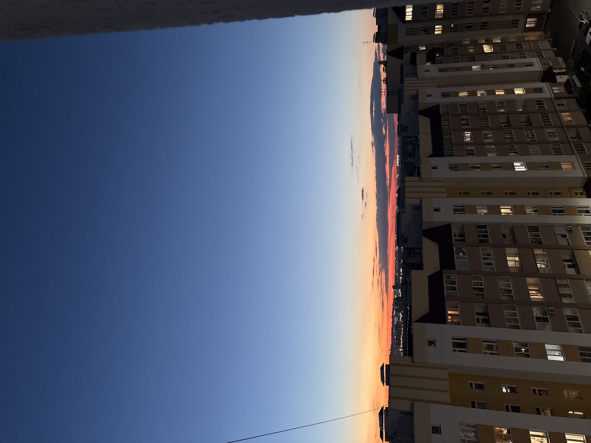 This is a vertically oriented, slightly tilted photograph of a twilight sky over a city. The sky is a gradient of deep blue at the top, transitioning to lighter blues and then to warm oranges and pinks near the horizon, indicating sunset or sunrise. A few wispy clouds are visible in the warmer toned part of the sky.
In the foreground and extending across the right side of the frame are multiple apartment buildings. These buildings are tall, multi-story structures with many windows. Many of the windows are illuminated, suggesting that people are inside and the evening is progressing. The architecture appears to be modern, with clean lines and a mix of light-colored facades, some with yellow accents. The buildings are tightly packed, hinting at an urban environment. A faint wire is visible stretching across the lower left portion of the image.
The scene conveys a sense of peaceful transition from day to night in a residential urban setting. The lighting suggests it is either early evening or late evening. No people are explicitly visible, but the illuminated windows imply human presence. There is no visible text in the image.