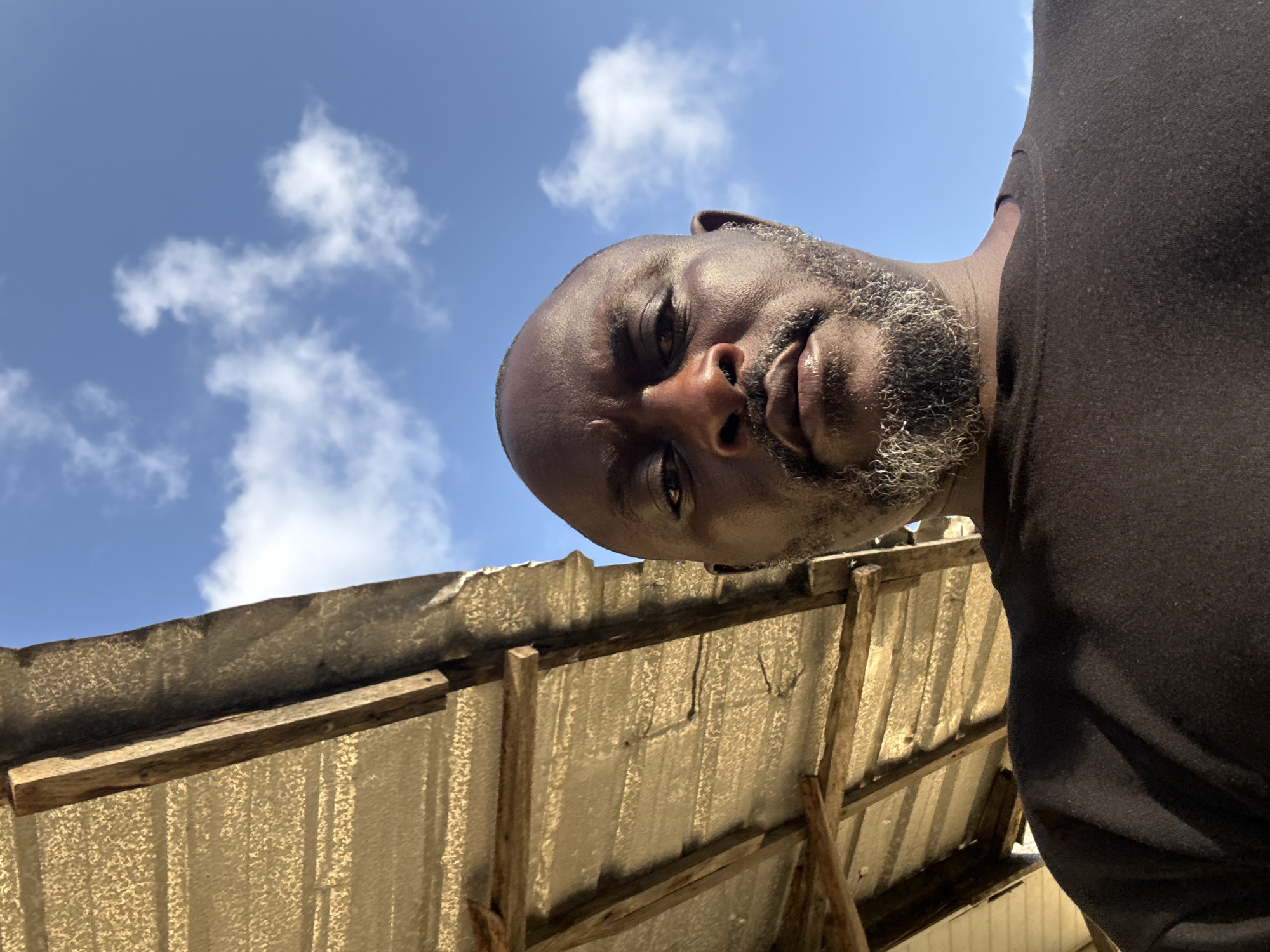 A low-angle, head-on photograph shows a man with dark skin, a shaved head, and a graying beard, positioned against a background of a bright blue sky with scattered white clouds. The man is wearing a dark-colored shirt. Beneath him is a corrugated metal roof supported by wooden beams. The location is inferred to be Ado, Nigeria. The scene is outdoors in daylight. The man is looking down toward the camera.