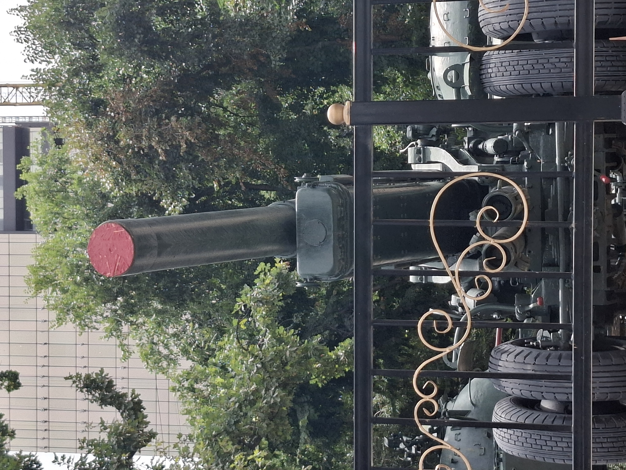 The image shows a large, dark green artillery piece with a red cap on its barrel, positioned behind a black metal fence with decorative gold scrollwork. The artillery piece has large tires and appears to be a static display. In the background, there are trees and a modern building with a grid-like facade, suggesting an urban environment. The overall scene is a clear, well-lit daytime shot. This artillery piece is located in Almaty, Kazakhstan, specifically at the Museum of the Armed Forces, which is located in Panfilov Park.