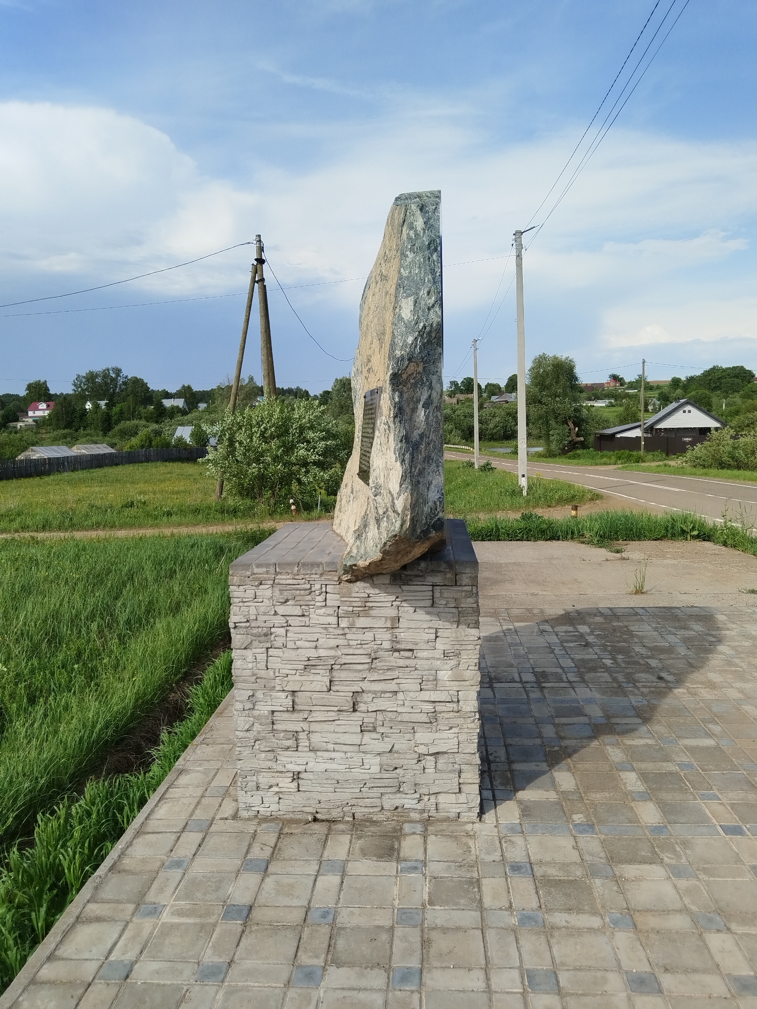 The image depicts a stone monument situated in Чумна, Russia. The monument is constructed with a base of layered gray stones and a tall, roughly hewn, slab of greenish-gray rock affixed on top. A plaque is visible on the rock slab. The monument is positioned near a paved area made of patterned gray tiles. To the left, there is a grassy field, and in the background, a road runs alongside power poles. Houses and trees are visible in the distance, underneath a sky with scattered clouds.