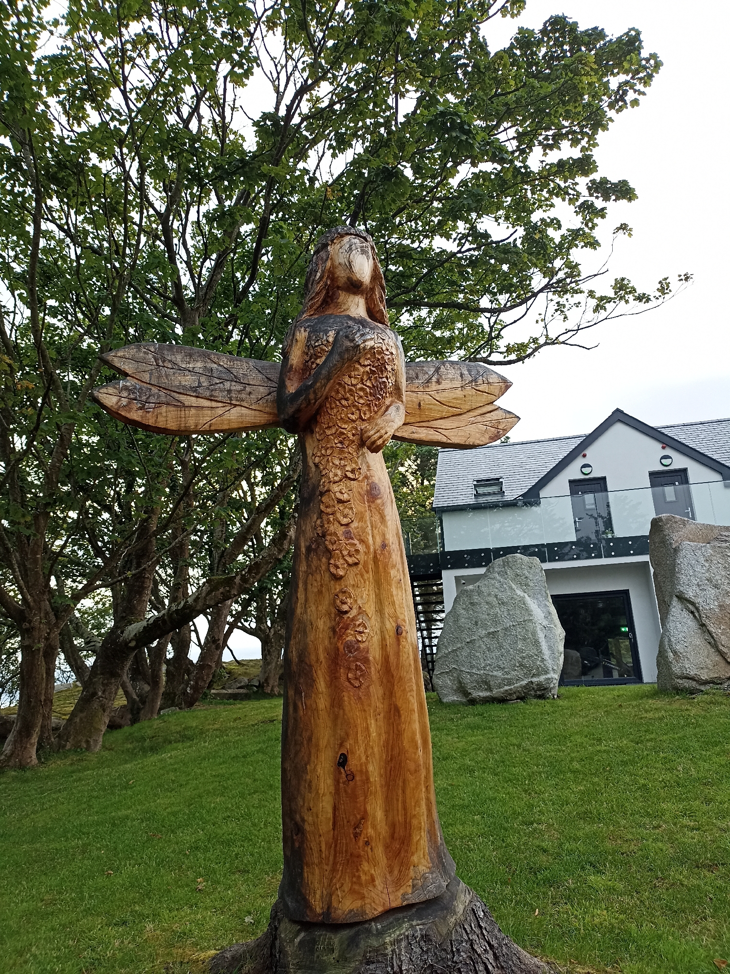 The image captures a wooden sculpture of a winged figure standing in a grassy area. The sculpture is carved from a light-colored wood and features wings on either side of its torso, which is adorned with a floral pattern. The figure's head is tilted slightly upward, and it appears to be wearing a crown. In the background, a modern house with a grey roof and white walls is visible, along with large rocks and trees. The scene is set in a slightly overcast outdoor environment.