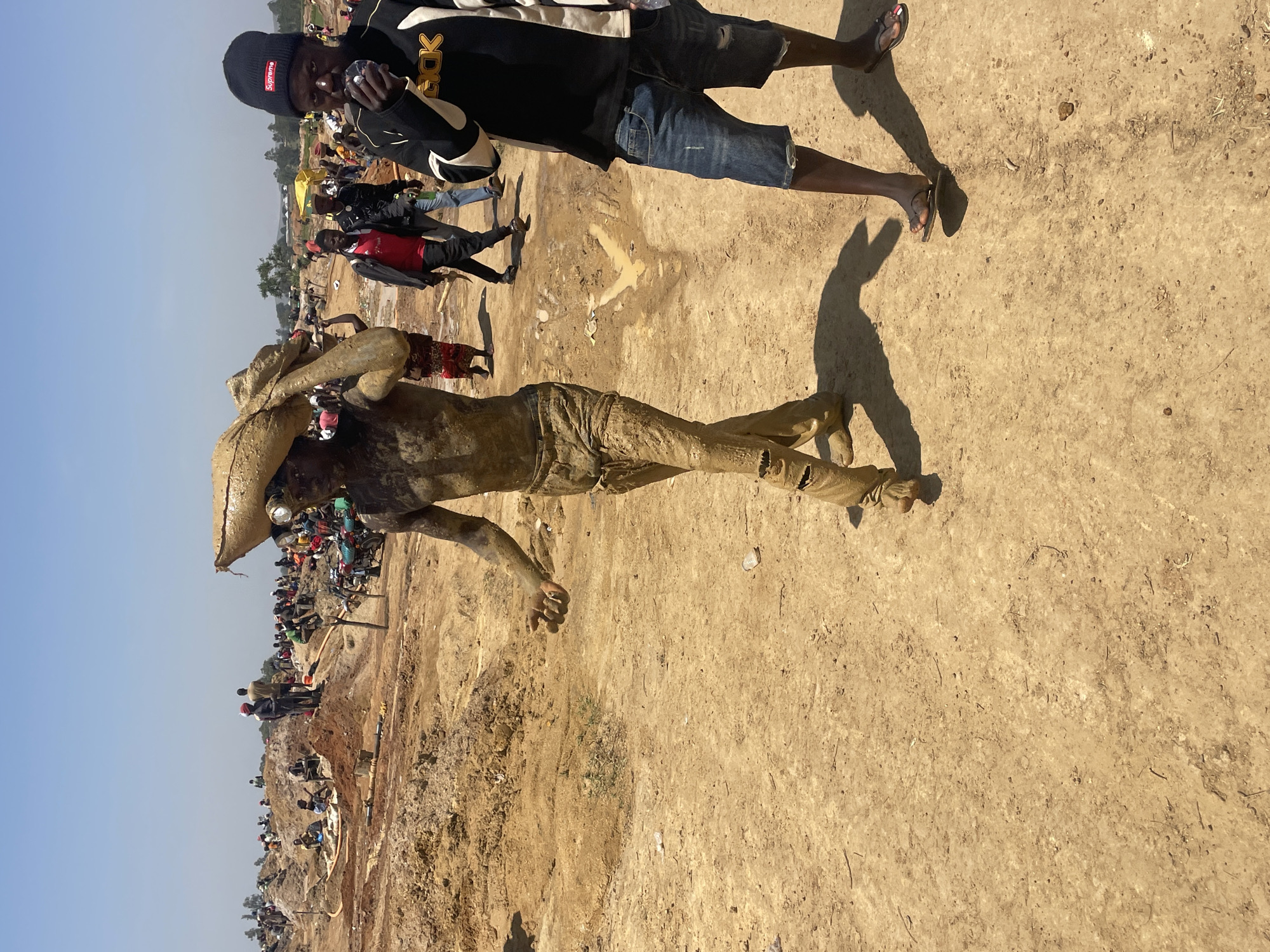 In Maiyanga, Nigeria, a wide-angle shot shows an open-pit mining operation under a clear, blue sky. In the foreground, a man covered in mud lies prone, holding a mud-covered sack over his head. A second man stands beside him, holding a camera. The second man wears a black and white jacket, a black "Supreme" beanie, and denim shorts. Both men are dark-skinned. In the background, a large crowd of people work in the muddy, excavated landscape. Hoses and digging equipment are visible. The ground is mostly bare dirt and mud.