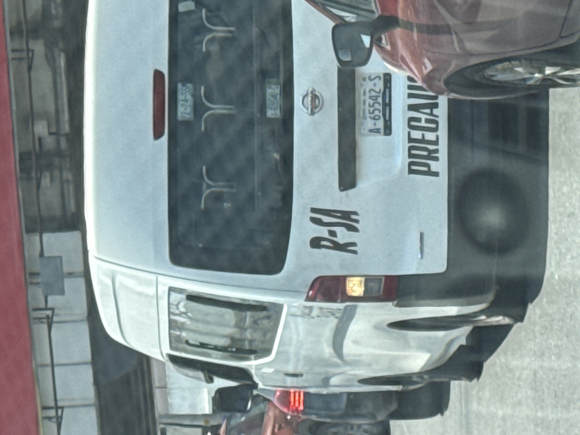 The image depicts the rear of a white Nissan NV350 van in Puebla, Mexico. The license plate reads "A-65542-S." The van has the words "PRECAUCIÓN" and "R-SA" vertically printed on the back. The vehicle is positioned next to a red car in what appears to be a city street environment. The van has a Nissan logo on the back and additional plates are visible on the rear window, with “72-24-55” discernible on one.
