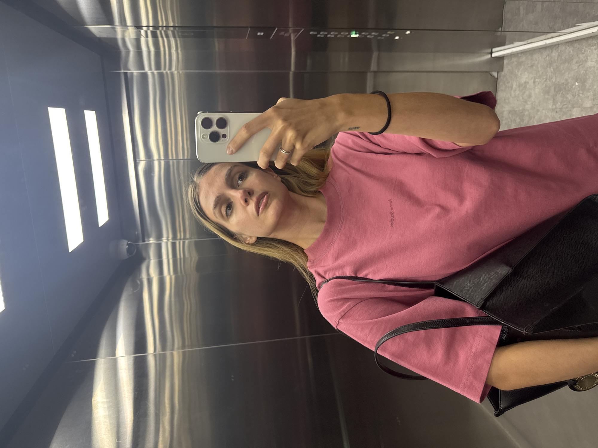 A woman takes a selfie in an elevator with mirrored walls. She is wearing a pink t-shirt and has a black bag slung over her shoulder. She is holding a white smartphone in her hand, taking the picture in the reflection. The elevator has metal walls and a row of buttons at the top. The floor appears to be light grey. The city and specific location are unknown.