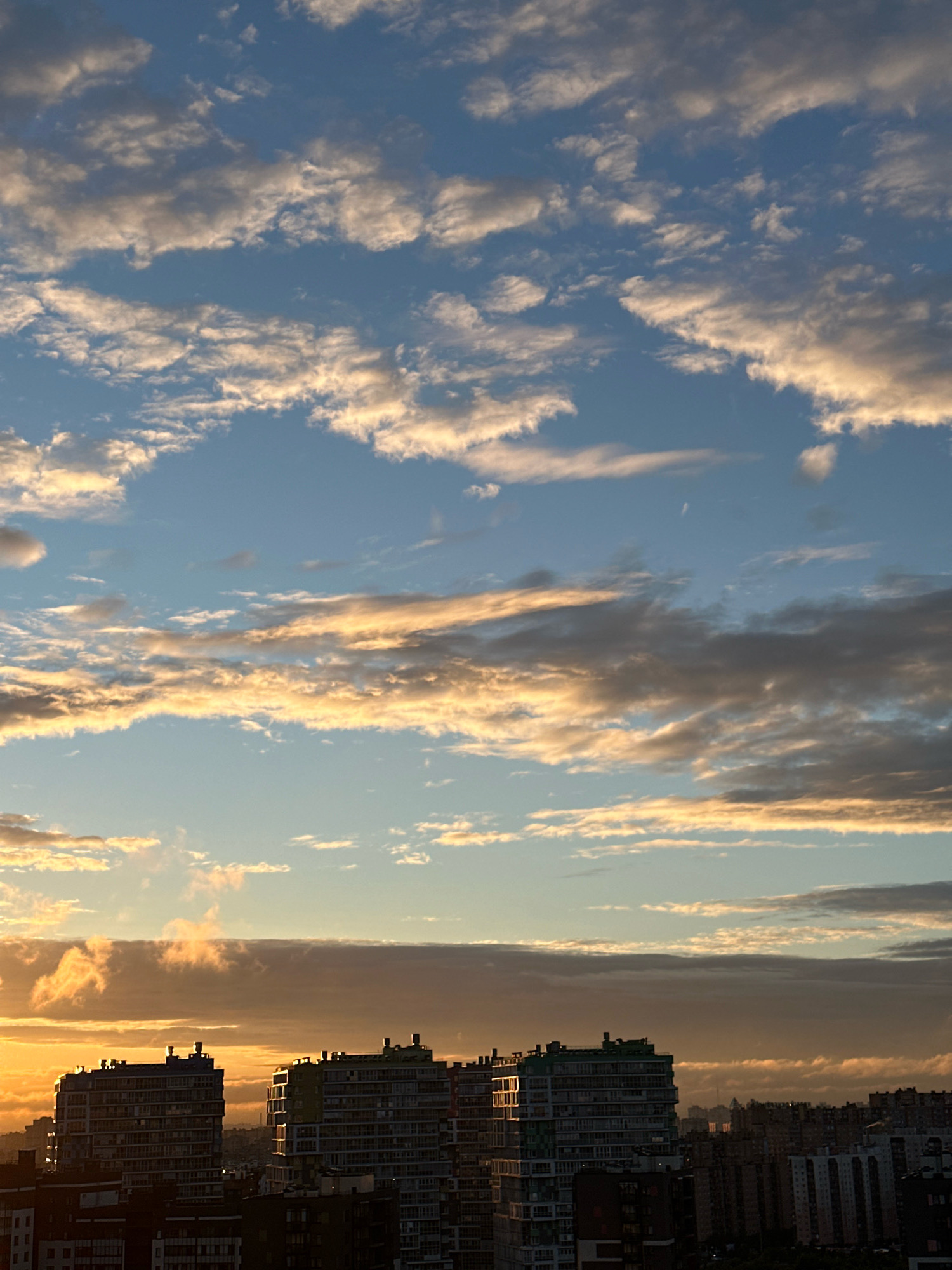 The image shows a cityscape at sunset or sunrise, with several multi-story buildings silhouetted against a sky filled with clouds. The clouds are illuminated from below by the warm glow of the sun, creating streaks of orange and yellow. The buildings appear to be apartment complexes, with many windows visible. Some of the buildings are taller and more modern than others, which appear older and more densely packed. The sky is a gradient of blue, transitioning to orange and yellow near the horizon. There is no visible indication of people or specific actions. The location is identified as Novaya Pustosh, Russia, suggesting a residential area in this city.