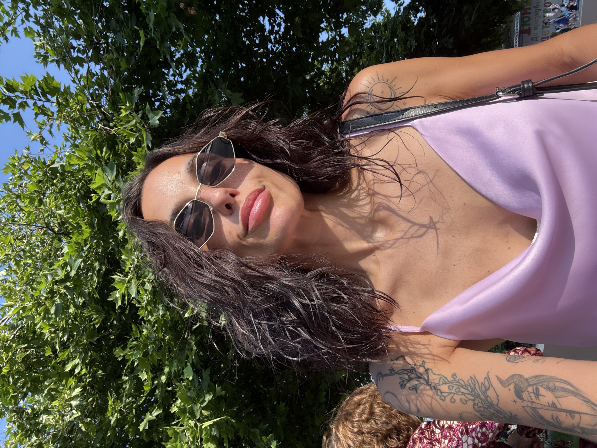 A female individual is depicted in an outdoor selfie, illuminated by direct sunlight which casts dappled shadows from overhead foliage onto her skin. She has dark, wet-styled, shoulder-length hair and is wearing dark, gold-framed, hexagonal sunglasses. Her lips are full, glossy, and slightly puckered. She wears a light lavender satin camisole with a cowl neck, and a black strap from a bag is visible over her right shoulder. Visible tattoos include a complex floral/vine and an inverted horned face design on her left bicep/forearm, and a crescent moon within a radiant sun on her right shoulder. To her left, the back of another person's head and shoulder, featuring curly, light brown hair and a patterned red top, is partially visible. The background is dominated by lush green foliage of large trees and patches of clear blue sky. In the far right background, a white banner with Cyrillic text, including "ЮНИ" (June) and "детс" (children's), is visible, suggesting a public event or festival. This linguistic and contextual evidence indicates the scene is located in an outdoor public space, likely a park or square, in the city of Pleven, Bulgaria.