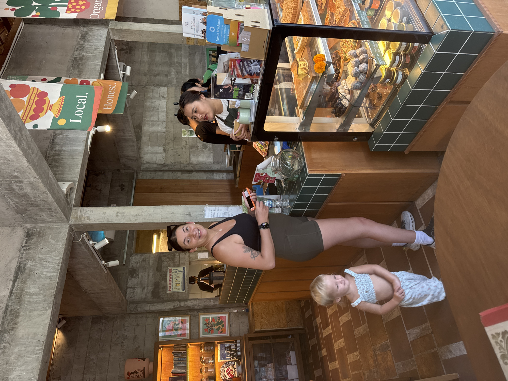 In a cafe in Mengwi, Indonesia, a woman and a young child stand near a counter displaying pastries. The woman, wearing a black tank top and olive-green shorts, holds a smartphone and a small bottle. The child, in a floral two-piece outfit, looks down. Behind them, a cafe employee serves customers at the counter. The cafe has a rustic interior with concrete walls and shelves stocked with books and jars. Banners with "Local." and "Honest." are hung from the ceiling. The display case features a variety of desserts, including cakes and chocolates.