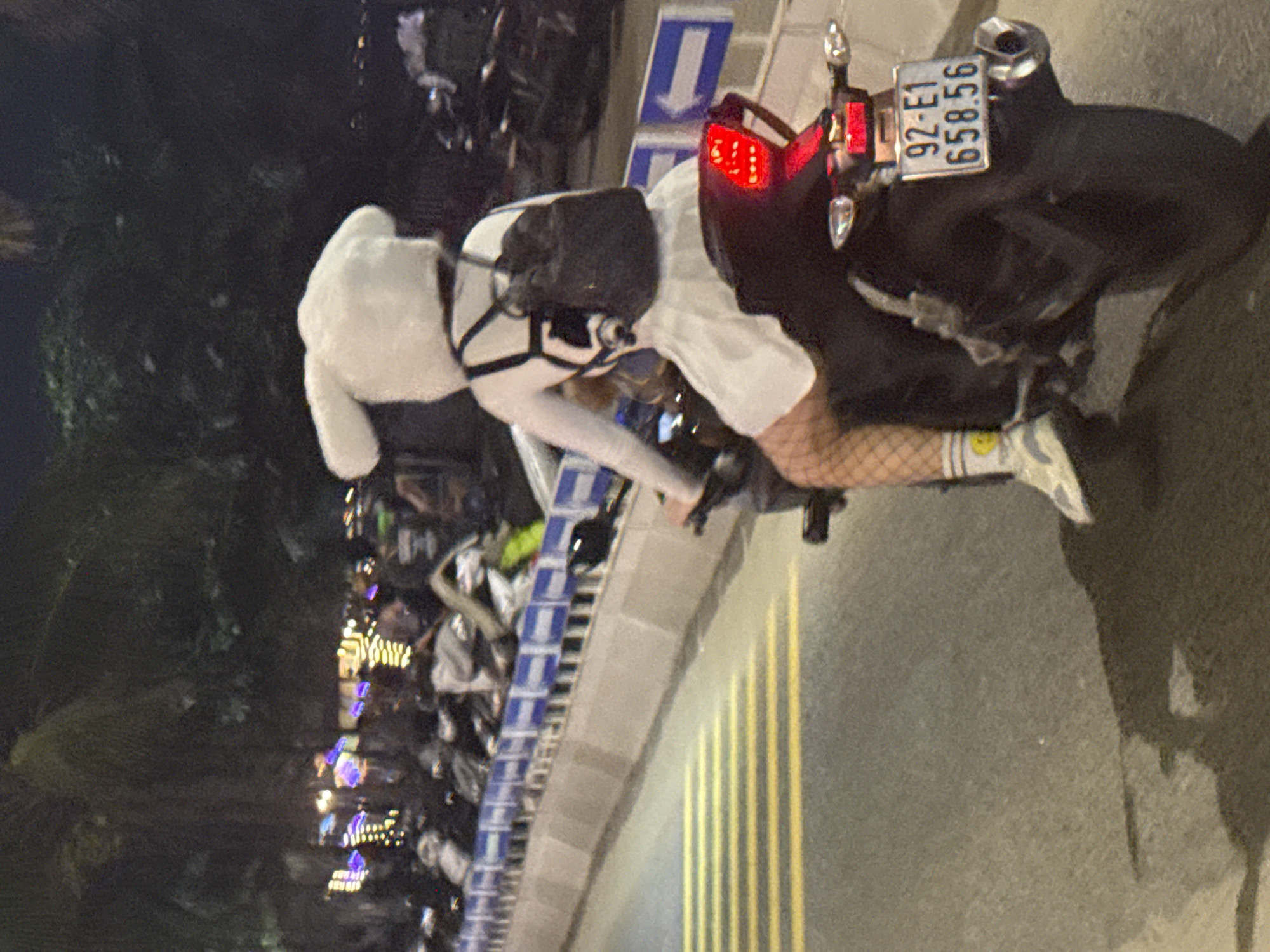 The image captures a scene at night. A person is seen on a motorcycle with license plate "92-E1 658.56" at roadside in the Vietnamese city of Đà Nẵng. The person is dressed in white clothing and is seen in partial view. The street is marked with yellow lines. The location appears to be at the corner of Trần Phú and Bạch Đằng Street, Đà Nẵng, near the area by the bridge. In the background, there is a crowd of people by the roadside and some foliage. The lighting indicates it is nighttime. The motorcycle is near a roadside curb.