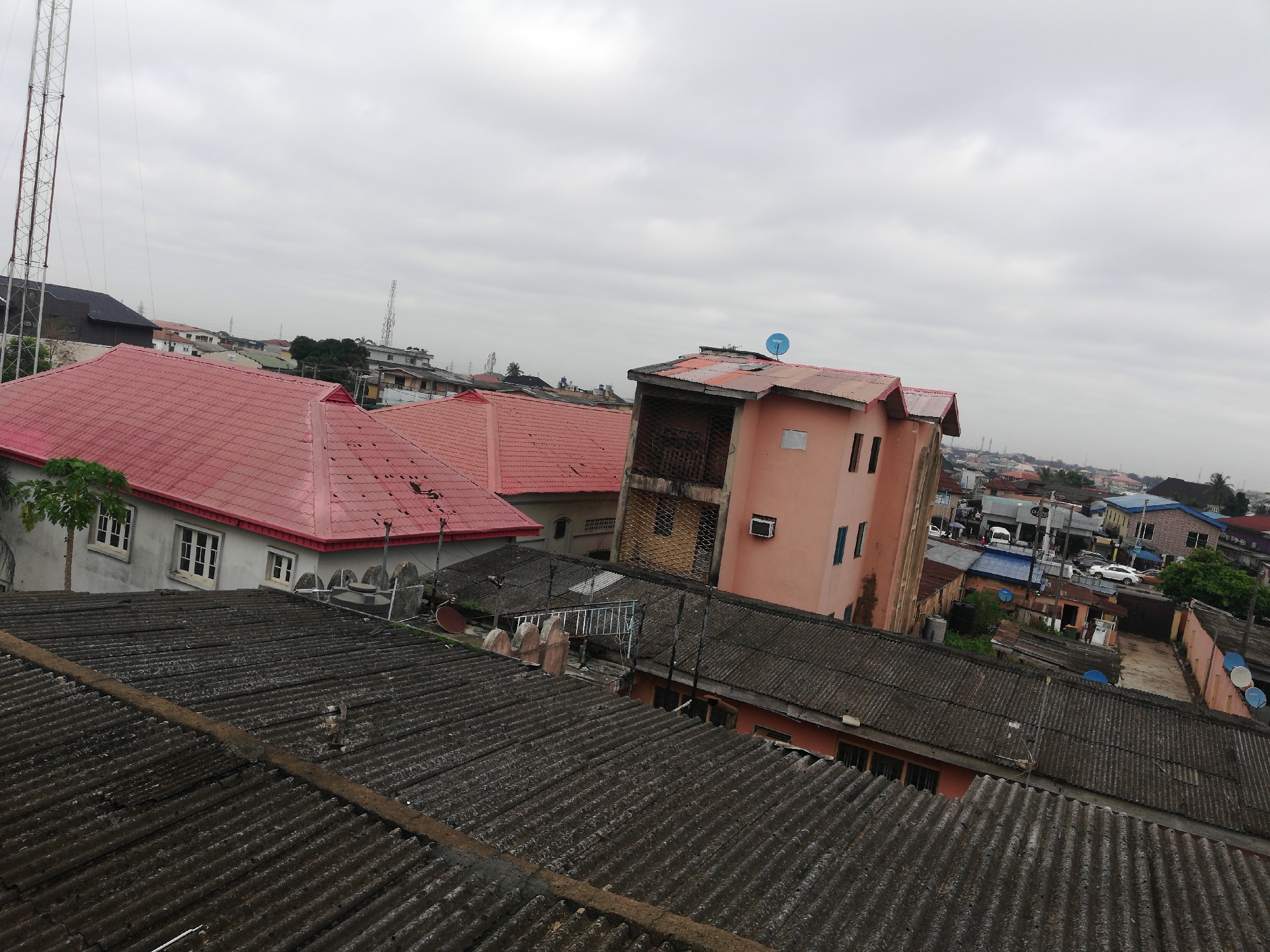 The image captures an elevated view of a dense urban environment, presumably a residential area in Unknown, Unknown. The foreground is dominated by a corrugated rooftop. In the midground, structures include buildings with red and grey corrugated roofing, a light-pink building featuring what appears to be an air-conditioning unit and blue satellite dishes. A tall telecommunications tower stands in the background, adding to the urban feel of the scene. The sky is overcast, suggesting cloudy weather conditions. Vehicles are visible in the streets below, implying a moderate level of activity in the area.
