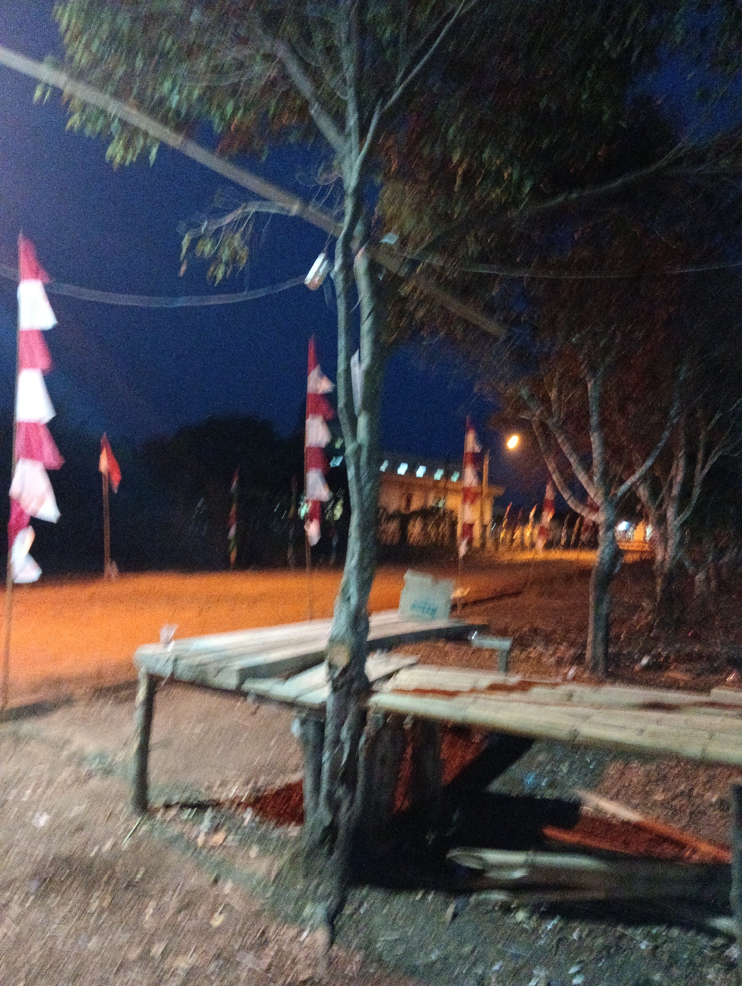 The image depicts an outdoor scene at night in Pematang Jaya, Indonesia. A tree stands in the foreground, its branches casting shadows. In the background, several Indonesian flags, with red and white horizontal stripes, are planted on poles. A wooden structure, possibly a bench or table, is visible in the mid-ground. Further in the distance, buildings with illuminated windows are present. The ground appears to be dirt or gravel. The lighting suggests it is twilight or early night.