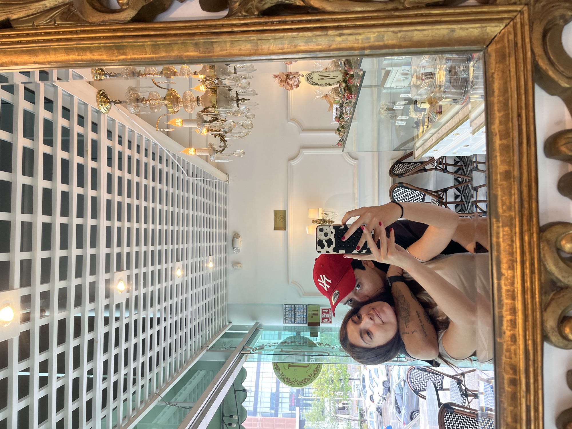 A mirror selfie captures two individuals, likely a couple, posing in what appears to be an upscale cafe or restaurant in San Bernardino Tlaxcalancingo, Mexico. The image is taken in a large, ornate gold-framed mirror, reflecting both the people and their surroundings.
The person closest to the camera, with long dark hair and wearing a light-colored top, is taking the photo with a smartphone featuring a distinctive black and white cow-print case. This person has red nail polish and a tattoo on their right arm, which includes some indecipherable text. Peeking over their shoulder is another individual wearing a red baseball cap with the white "NY" (New York Yankees) logo and a dark top. Both individuals appear relaxed, with faint smiles.
The interior reflected in the mirror reveals a bright, elegant setting. A large, ornate gold chandelier with multiple glowing lights hangs from the ceiling. The walls are white, featuring classic decorative molding. A white latticework partition or wall is visible on the left side of the reflection. In the background, there's a display case filled with what looks like pastries or decorative items, hinting at a bakery or dessert shop. Bistro-style chairs with distinctive black and white patterned seats and backs are also visible, suggesting a seating area.
Through large windows reflected in the mirror, an urban street scene is visible outside. Parked cars line the street, and green trees and multi-story buildings can be seen under a bright, clear daytime sky. Several small signs are visible on the interior walls and reflected in the windows, but any text on them is illegible due to distance and reflection.