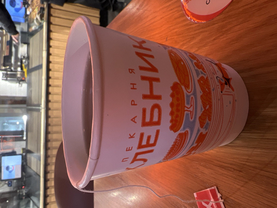 The image depicts an interior scene within a "Khlebnik" bakery, a chain operating in Saint Petersburg, Russia. A white paper cup, approximately 12-16 ounces, is centrally positioned on a light brown wooden table in the foreground. The cup contains a dark liquid, consistent with brewed tea, from which a white string and a partially visible red tea tag dangle over the left side. The cup's exterior features the Cyrillic text "ПЕКАРНЯ ХЛЕБНИК" (Bakery Khlebnik) in red-orange letters, above a decorative band containing illustrations of various baked goods like pies and croissants, and a small red stylized dog. To the immediate right of the cup, a small, partially visible red object, possibly heart-shaped or circular with white text, rests on the table. In the blurred background, a commercial counter area with glass display cases is visible. A person in dark clothing is partially observed behind the counter, appearing to operate a piece of industrial-style beverage or food preparation equipment. Two electronic display screens are mounted above the counter area. The scene is illuminated by interior lighting, casting warm tones on the wooden surface.
