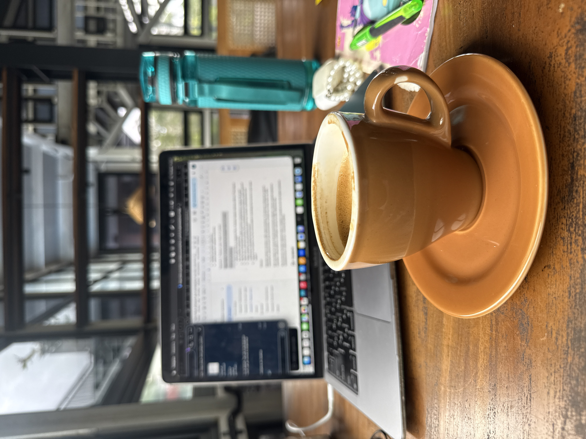 A low-angle, close-up shot of a brown espresso cup and saucer sits on a wooden table. The cup is filled with dark espresso, topped with a layer of crema. To the left of the cup, a silver laptop is open, displaying a document on its screen. To the right of the cup, a turquoise water bottle stands upright, next to a stack of pink and white papers. A string of pearls and a green pen are also visible on the papers. In the background, a blurred view of a modern interior with metal beams and stairs can be seen, suggesting an indoor workspace or cafe, possibly in Kuta Selatan, Indonesia. The lighting is natural, coming from an unseen source, creating soft shadows on the wooden surface.