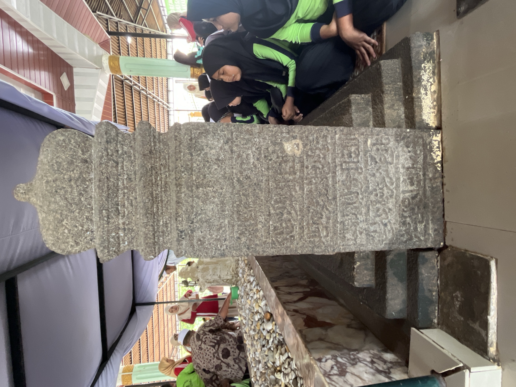 In Beuringen, Indonesia, a group of people are gathered around a large, ornate stone monument, possibly a tomb or historical marker. The monument is carved with intricate inscriptions and features a stepped base. Several individuals, including women in hijabs and a man in traditional attire, are present. Some are observing the monument, while others appear to be participating in a ritual, as indicated by a person pouring liquid from a green container onto the ground near the monument. The scene is set outdoors, under a large canopy or tent structure. The surrounding environment includes a tiled floor and a wall with marble-like detailing. The lighting suggests it is daytime, with sunlight filtering through the covered area.