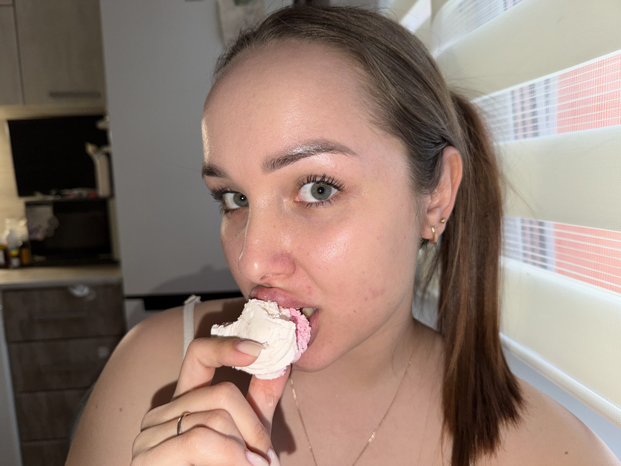 A woman is eating a meringue cookie indoors. She has fair skin, light brown hair pulled back into a ponytail, and green eyes. She is wearing a gold necklace and a gold ring. The meringue has a white outer layer and a pink inner layer. A kitchen with cabinets, a refrigerator, and other appliances is visible in the background. The woman is standing near a window with white and orange blinds. The location is not possible to determine.