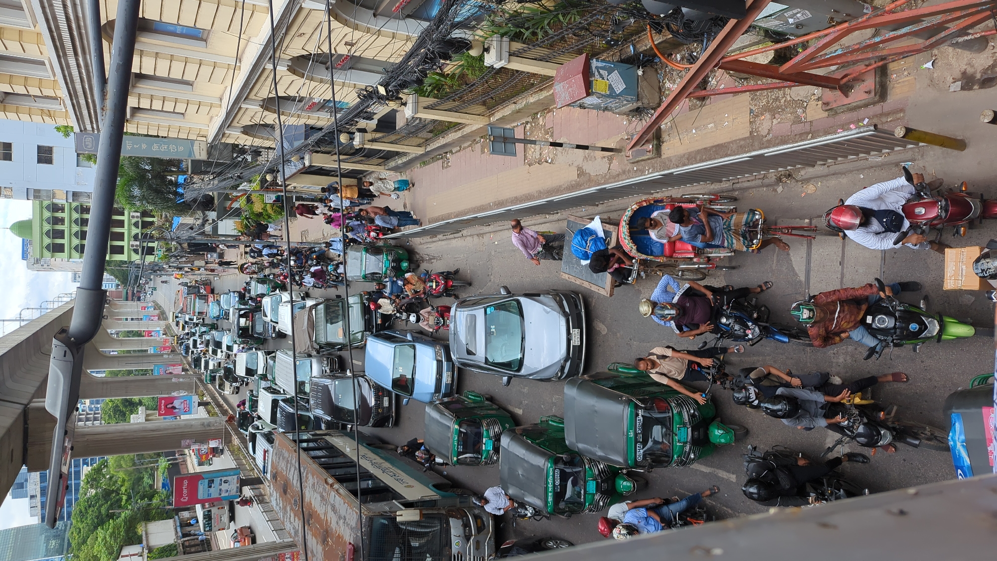 The image depicts a busy street scene in Dhaka, Bangladesh, likely during daylight hours, characterized by significant traffic congestion. A multi-lane road is filled with various vehicles including cars, buses, auto-rickshaws (CNGs), motorcycles, and a rickshaw. Several individuals are visible, many of whom are operating these vehicles or walking on the roadside.
In the foreground, a rusty-brown bus is parked on the left, partially obscuring the view of other vehicles. Ahead of it, a lighter colored bus is also present. Several green auto-rickshaws are prominent, with drivers and passengers inside. A light blue car and a grey sedan are among the cars in traffic. Motorcycles are numerous, with many riders wearing helmets. A pulled rickshaw with passengers is also visible.
Pedestrians are seen walking along the sidewalks and near the vehicles. The environment includes buildings lining the street, one with yellow facade and arched windows, and another further back with a green facade and dome. Overhead, a concrete elevated structure, likely part of a metro rail or flyover system, dominates the upper portion of the image, with supporting pillars. Overhead wires and streetlights are also present. The road surface appears to be asphalt, and the overall impression is one of urban density and heavy vehicular activity typical of Dhaka.