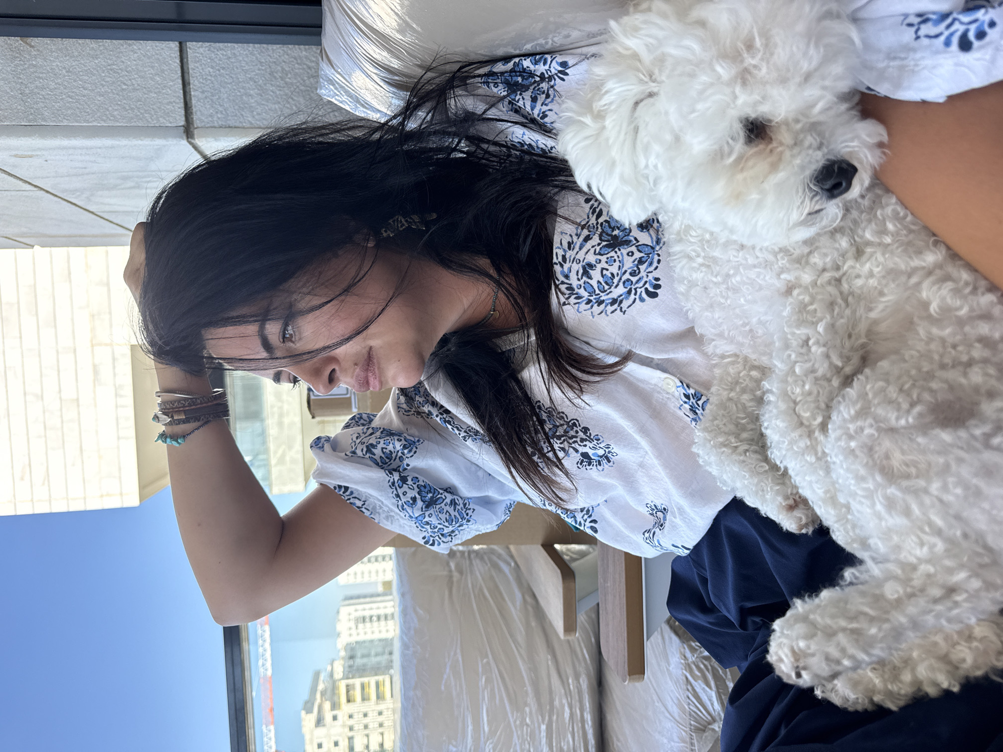 A medium shot shows a woman with dark hair and a white shirt with blue patterns. She is sitting on a couch with a white dog resting on her lap. The woman is looking to the left, with her right arm resting on her head. The dog appears to be sleeping. Behind the woman, through a window, is a view of buildings and a construction crane, indicating an urban setting. The scene suggests a casual, indoor/outdoor environment, likely a balcony or patio. The location is identified as Or Yehuda, Israel.