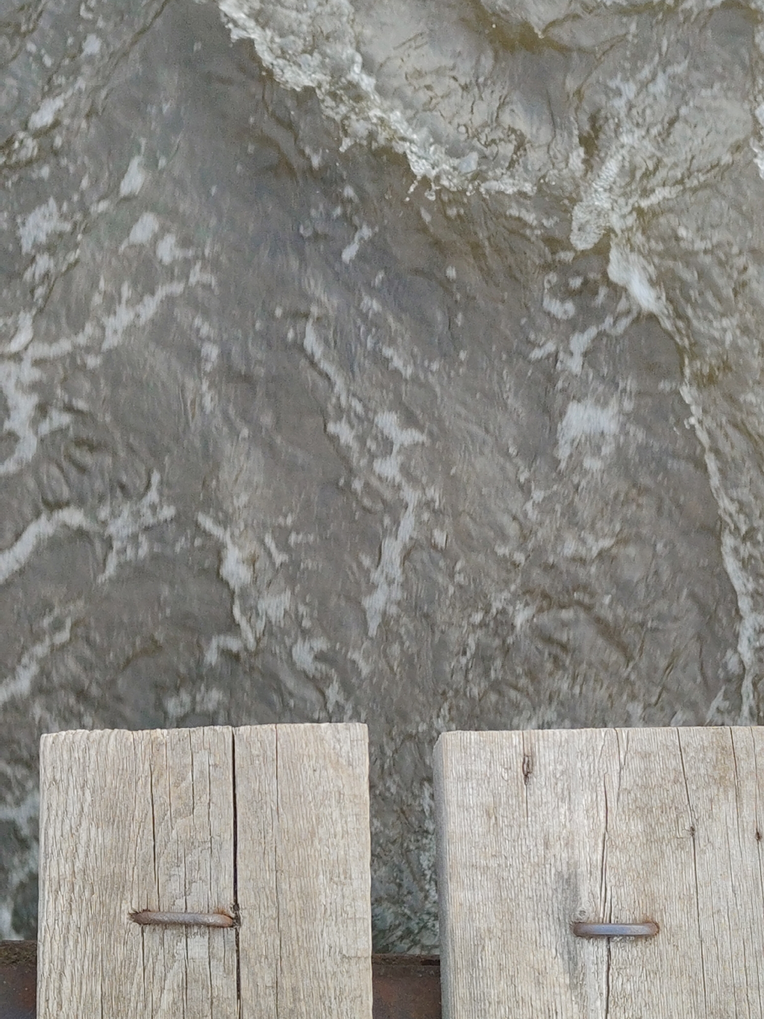 The image shows two weathered wooden planks in the foreground, secured with metal staples. Behind them is a body of water with turbulent, foamy surface, suggestive of a river or fast-moving stream. The location is unknown.