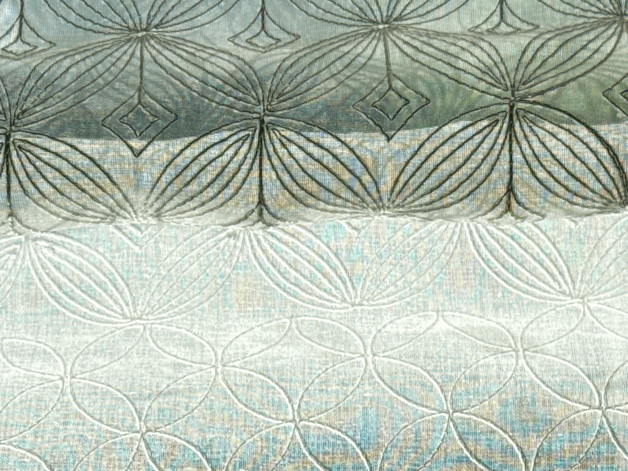 The image shows a close-up of patterned fabric. The pattern consists of repeating floral or leaf-like designs. The upper portion of the fabric has a darker, possibly embossed or embroidered, pattern with black lines, while the lower part features a lighter, possibly printed, pattern in white or light gray. The background color of the fabric appears to be a mixture of light blue, gray, and brown hues. The texture suggests a woven material. There is no information about the city of Milan, Italy, in the image.