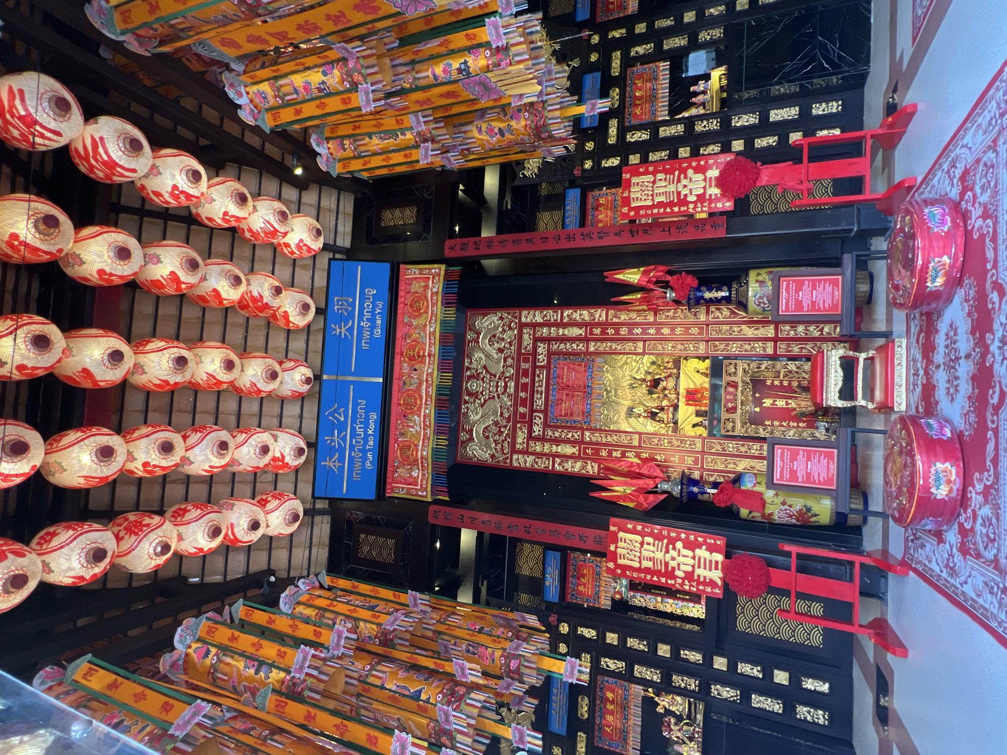 The image captures the interior of a temple located in Baan Hua Thanon, Thailand. The space is adorned with rows of red and white lanterns hanging from the ceiling, alongside yellow banners or pennants. The walls are intricately decorated with black and gold carvings, framed panels, and red signs with Chinese characters. A shrine is visible, featuring golden dragons and other ornate details, as well as statues and offerings. Signs in both Thai and Chinese identify the deities worshipped as Guan Yu and Pun Tao Kong. The floor is tiled and the overall lighting suggests it is either inside or under a covered structure. The space appears to be a place of religious observance, adorned with traditional Chinese decorations.