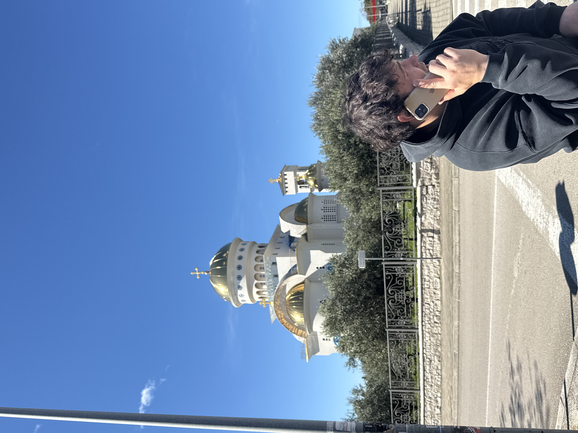 A medium shot depicts a man with curly dark hair, wearing a black hoodie, talking on an iPhone in a protective beige case. The man is positioned in the upper right of the frame, with the phone held to his ear. Behind him, a wrought iron fence separates him from a landscaped area with trees. The background showcases the Cathedral of St. John Vladimir in Bar, Montenegro, with its distinctive white walls and gilded domes. The sky is a clear, bright blue. The road, with its visible asphalt and painted lines, suggests an urban environment. A utility pole is at the bottom left.