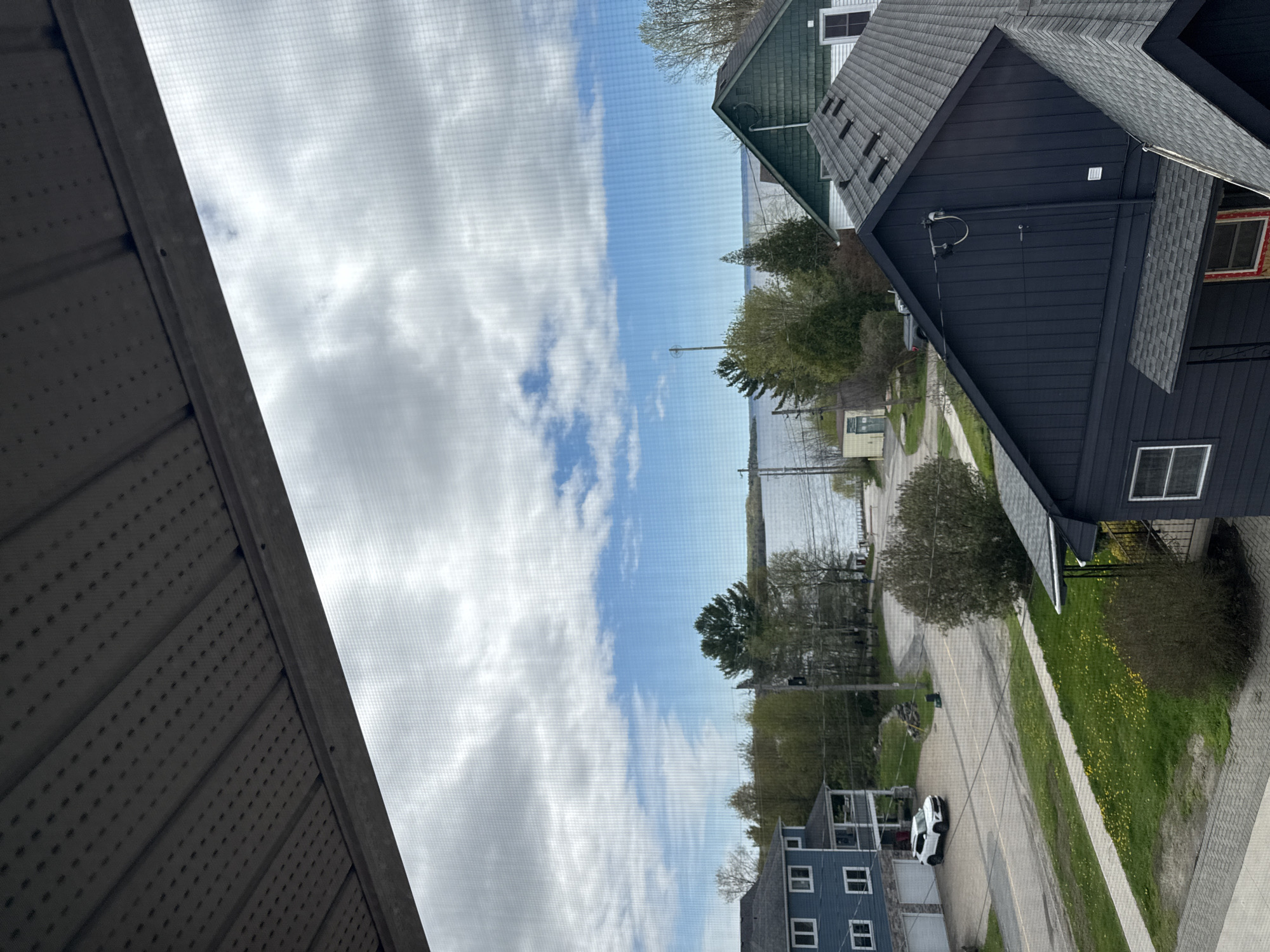 The image depicts an outdoor scene observed through a window screen. Visible in the background is a body of water with a shoreline dotted with trees and buildings. In the foreground, there are residential buildings, a street with a white car parked, green lawns, and various trees. The sky is predominantly blue with white clouds. The scene appears to be set in a residential area in a town or city of unknown location.