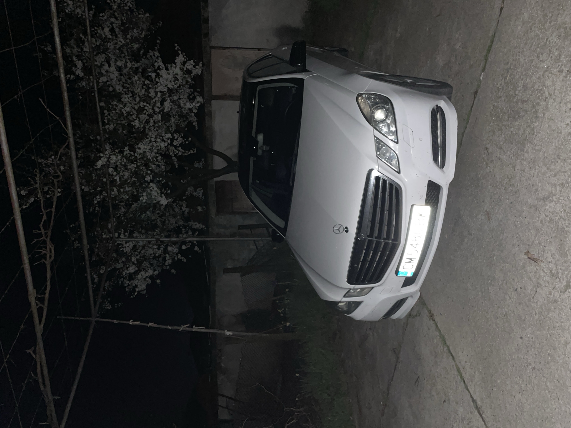A white Mercedes-Benz E-Class sedan is parked on a concrete driveway at night in Zlatograd, Bulgaria. The car is facing the camera. The license plate reads "CM 5487 BK". A flowering tree is visible on the left, behind a metal fence. The background is dark, with a partial view of a building. The scene is dimly lit.