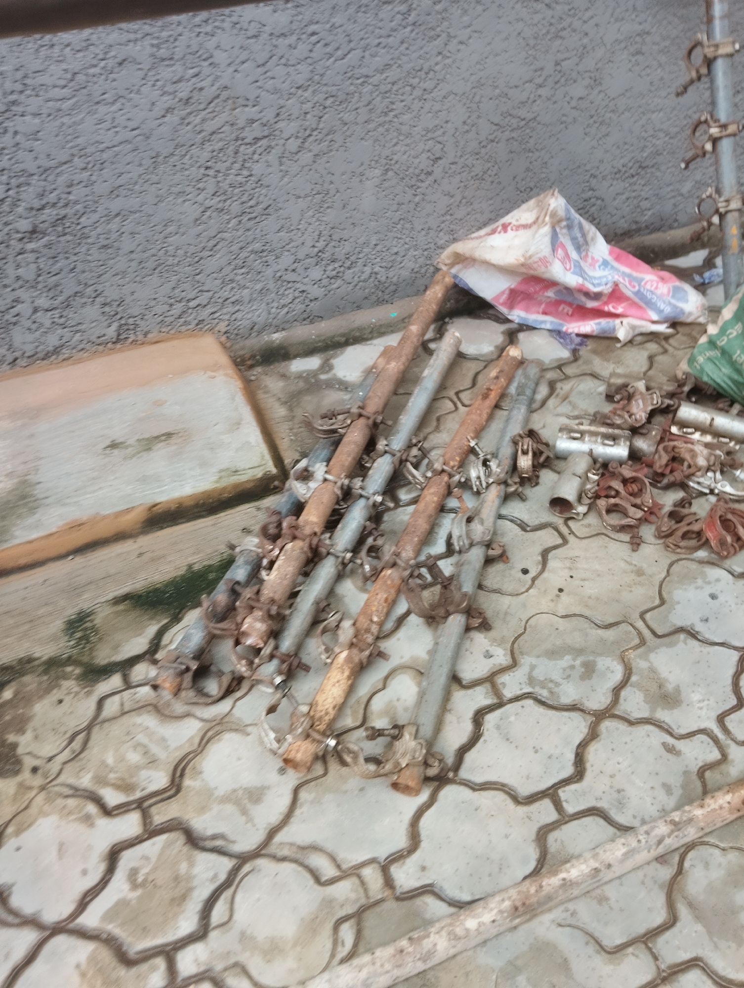 The image depicts a collection of scaffolding poles and clamps scattered on a patterned paved surface in front of a textured gray wall. The scaffolding poles appear to be aged and some show signs of rust. A partially opened, white and red striped bag, possibly containing construction materials, lies near the wall. To the left, a rectangular concrete slab is visible. The scene suggests an outdoor construction or storage area, consistent with locations found in various parts of Lagos, Nigeria. There are no people or discernible actions taking place in the image.