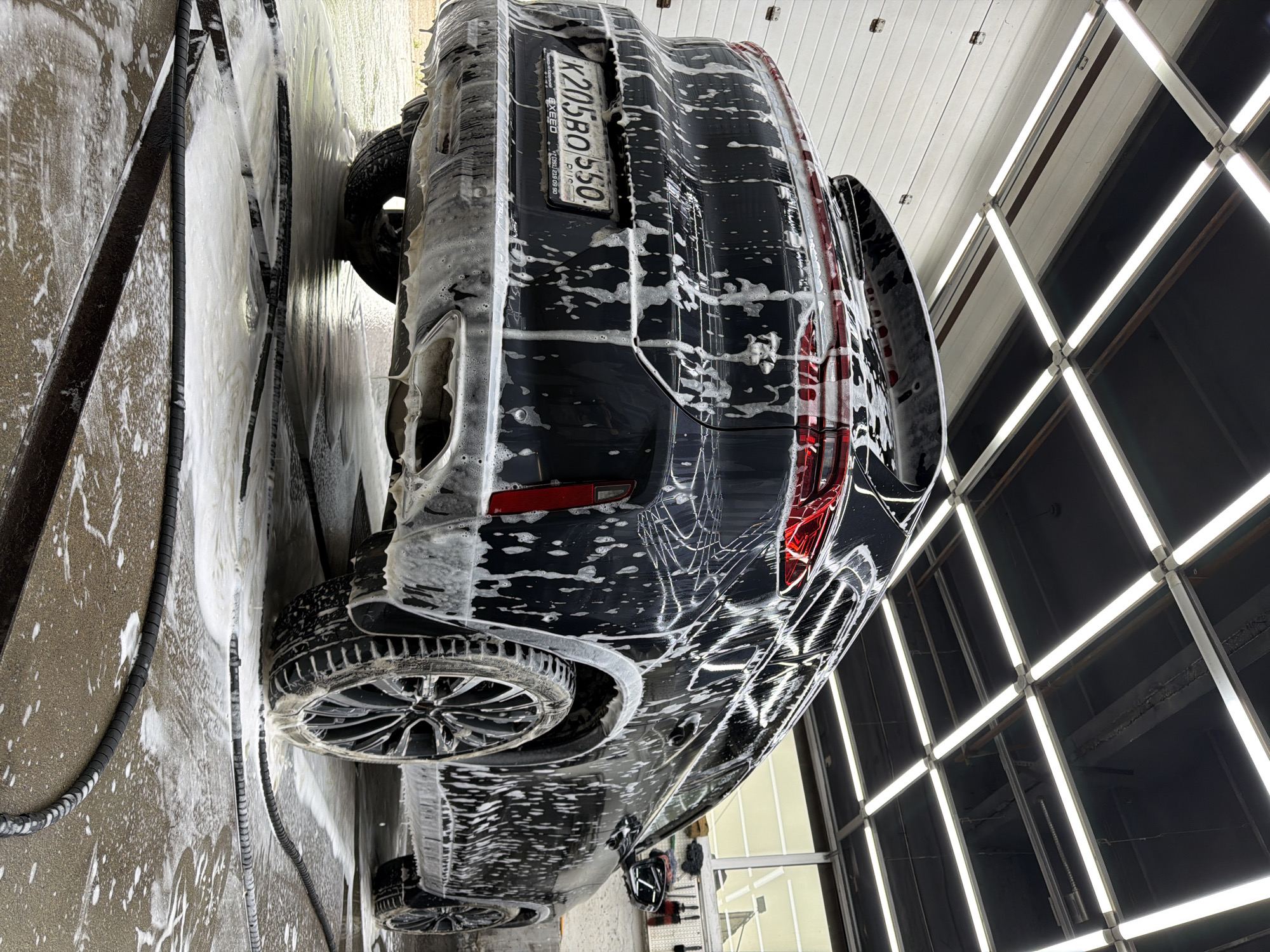A black SUV, covered in soap suds, is being washed in a car wash bay. The car is positioned at an angle, facing away from the camera, with its rear in full view. The license plate is visible and reads "K205BO 550". The car wash bay has a tiled floor with drainage channels, and a black hose lies on the floor, also covered in suds. Above the car, a grid of fluorescent lights illuminates the scene. The background shows a white corrugated metal wall and a large window, suggesting an indoor car wash facility. The image is taken from a low angle, emphasizing the car and its sudsy exterior. The location is identified as a car wash in Krasnoyarsk, Russia. There are no people visible in the image.